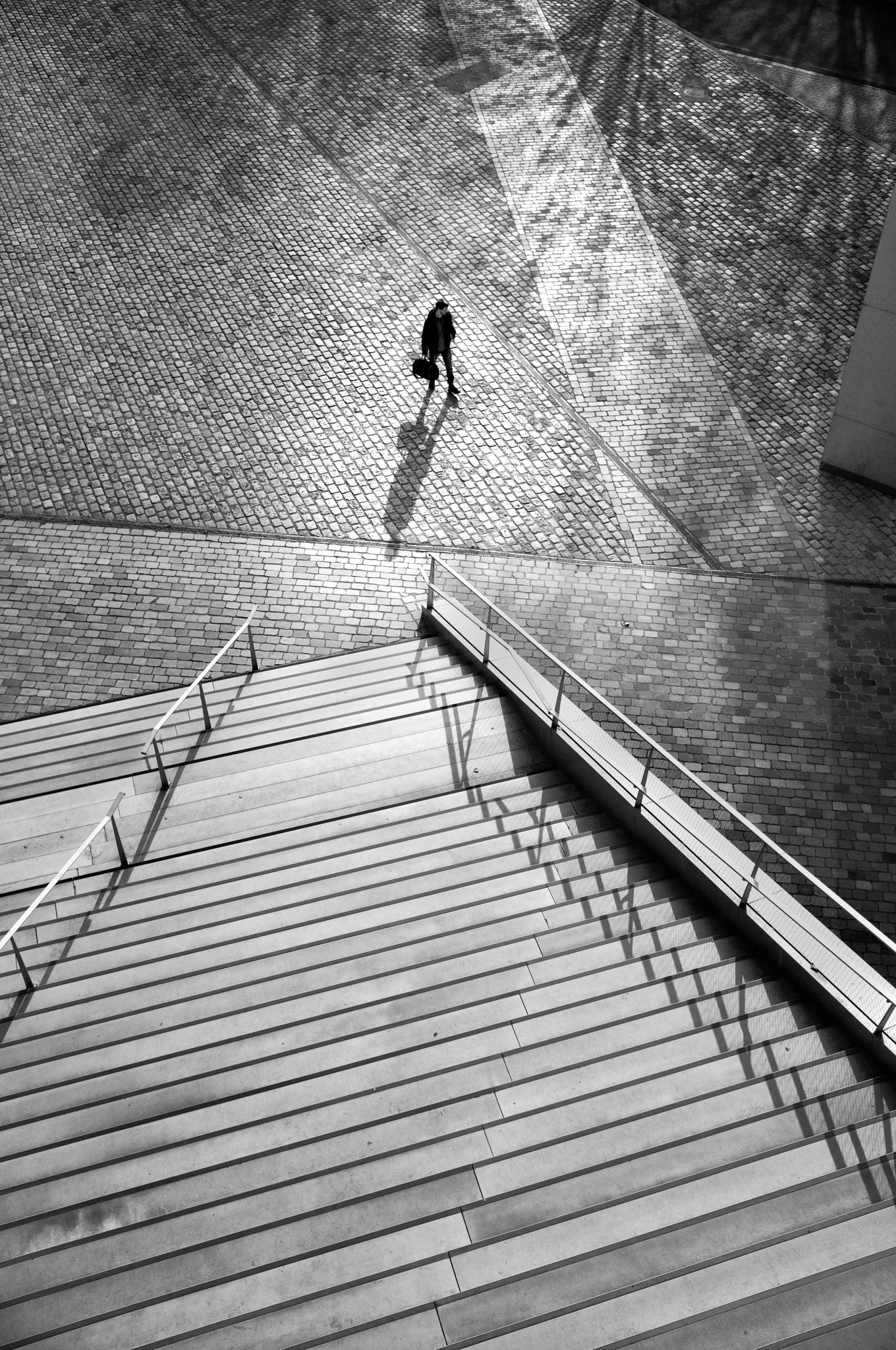 A person walking with a rolling suitcase on a cobblestone sidewalk, casting a long shadow in sunlight, viewed from an elevated perspective.