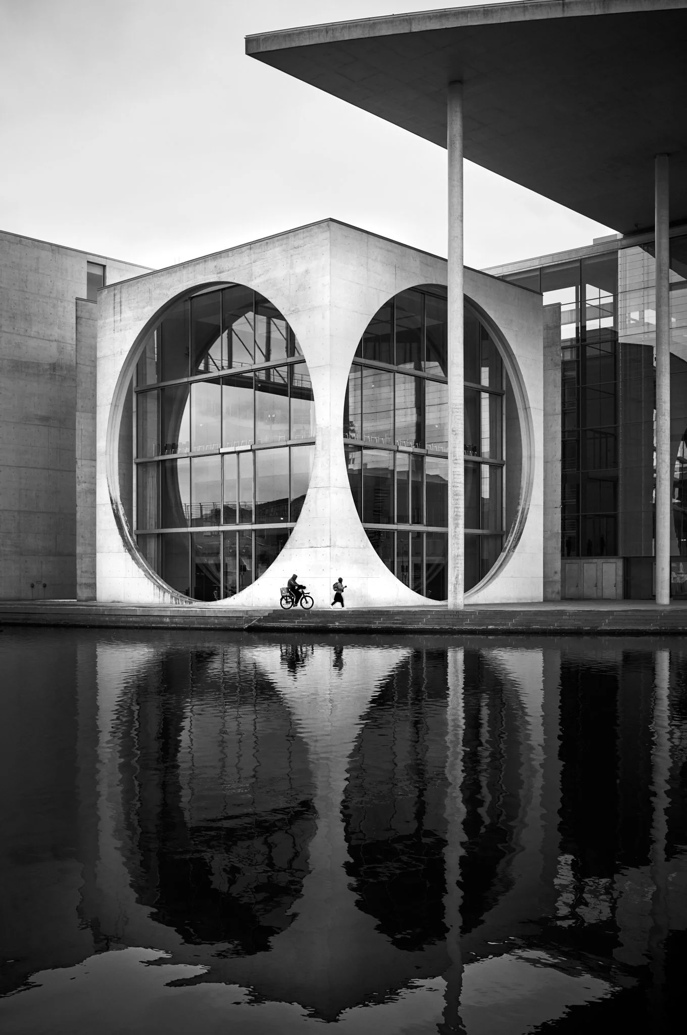 Modern building with large circular windows reflecting water and two people walking, one riding a bicycle, in black and white.
