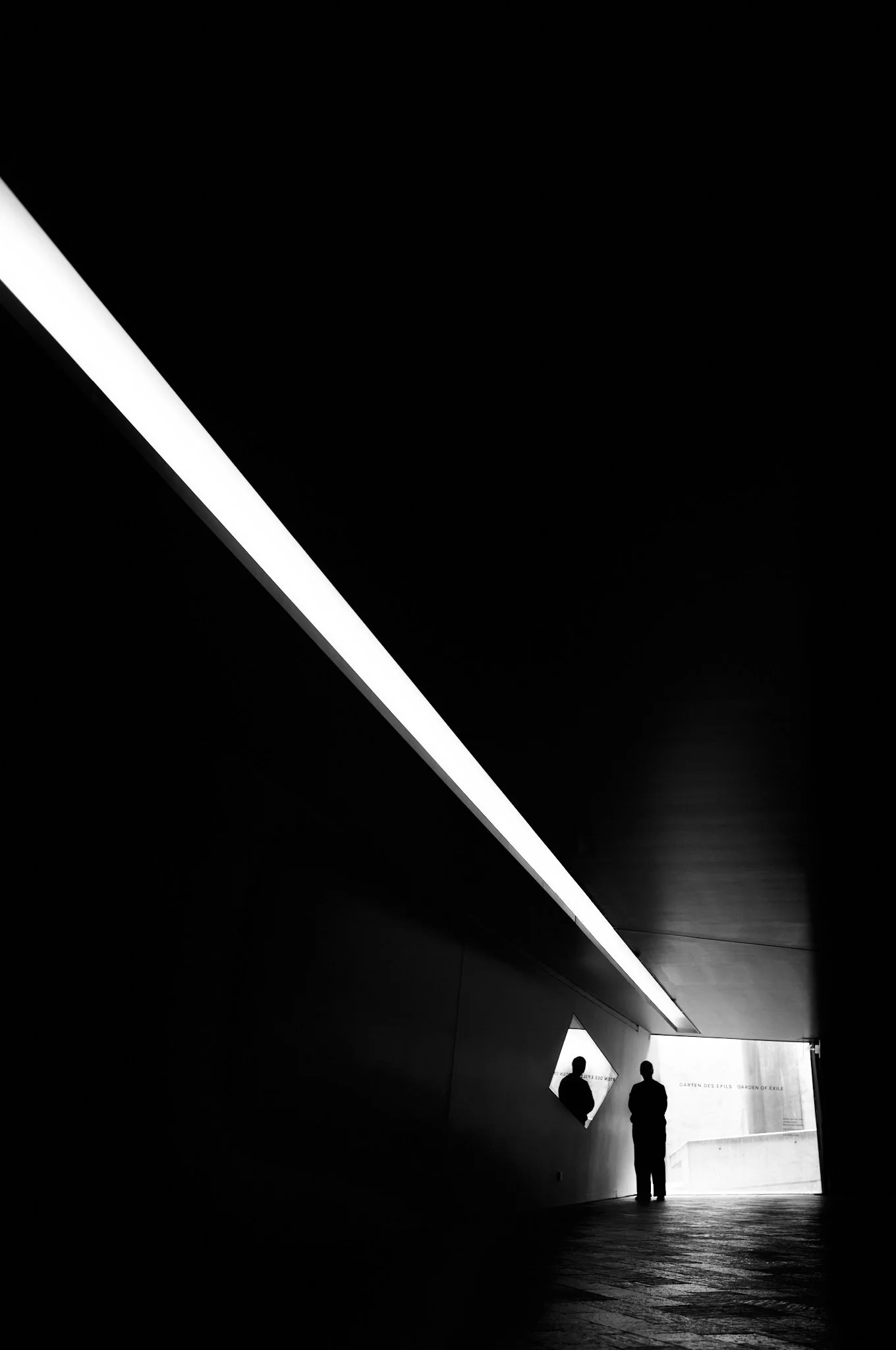 Silhouettes of two people standing beneath an overhang with bright light behind them in a dark urban setting.