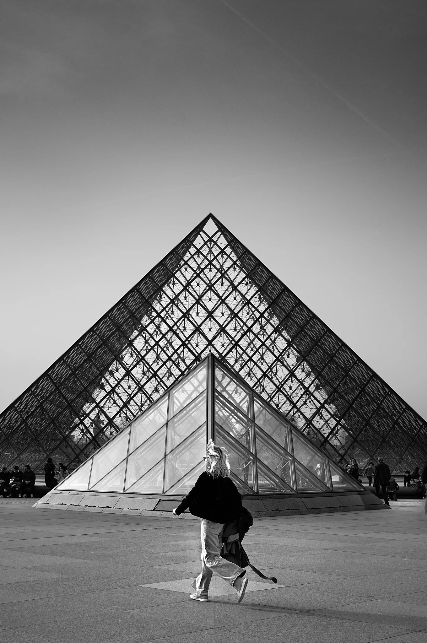 People walking and sitting in front of the Louvre Pyramid in Paris, France, with the glass and metal pyramid structure against a gray sky.