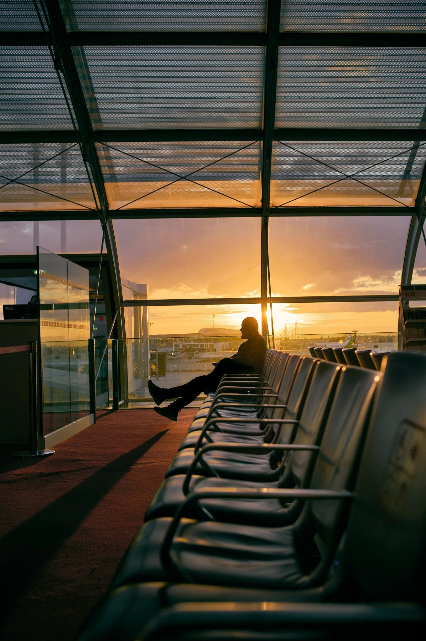 A person sitting alone in an airport terminal, facing the window during sunset, with empty chairs in the foreground and airplanes visible outside.