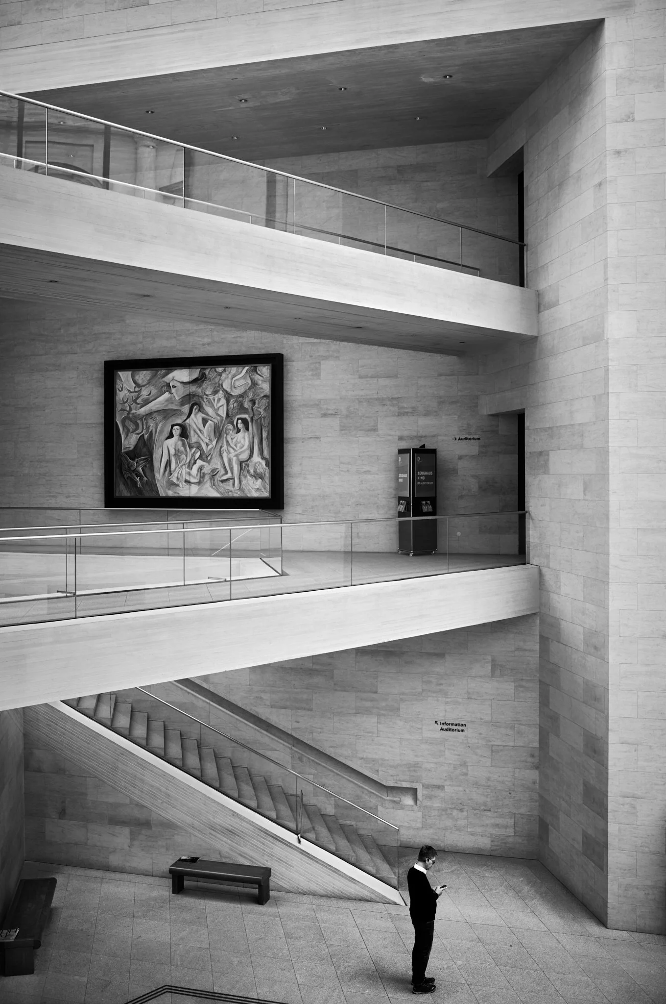 Interior view of a modern museum or gallery with curved walls, featuring an abstract painting on the wall, a staircase, and a man wearing a face mask looking at his phone.