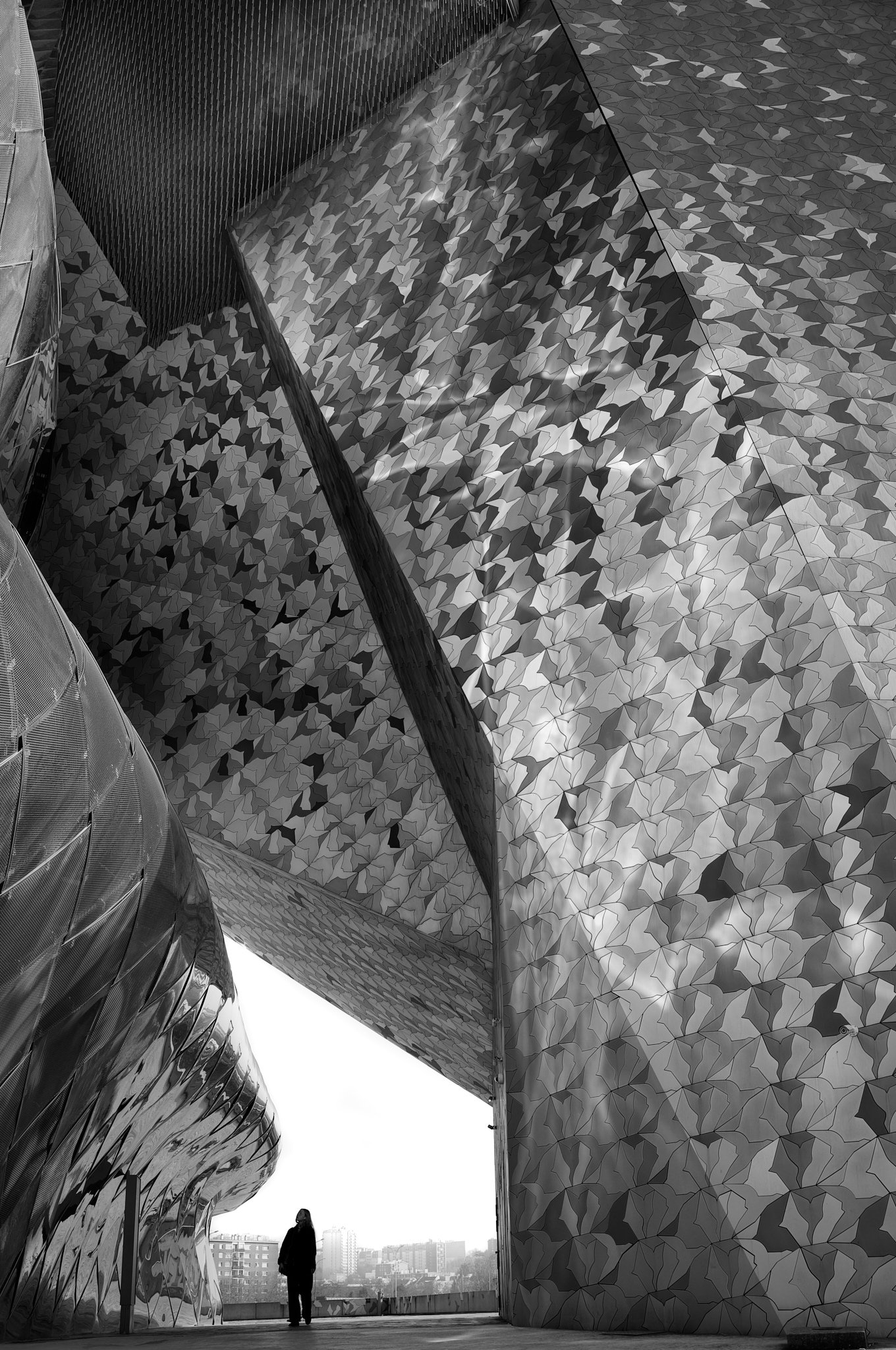 A black and white photo of a person standing under an architectural structure with curved, metallic and mosaic patterns on the walls.