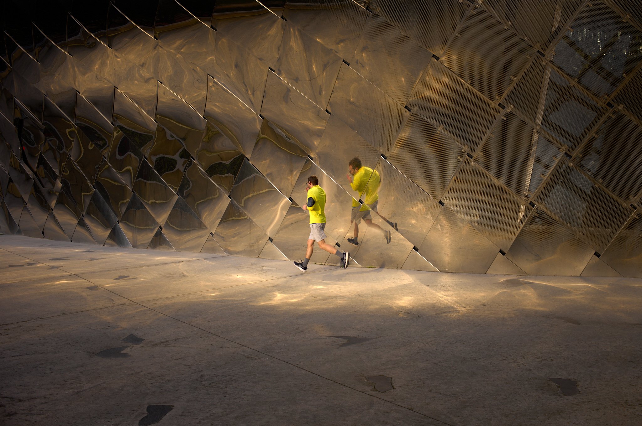 Two young men in yellow shirts and gray shorts walking in front of a modern, reflective metallic architectural structure. The structure has an angular, geometric design with shiny surfaces that mirror the surroundings.