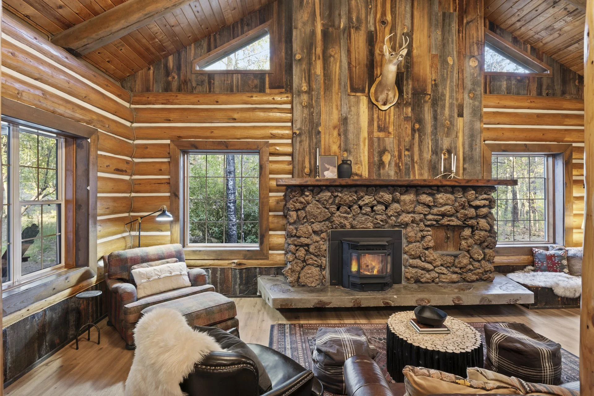 Interior of a rustic log cabin living room with a stone fireplace, wood-paneled walls, and large windows showing trees outside.