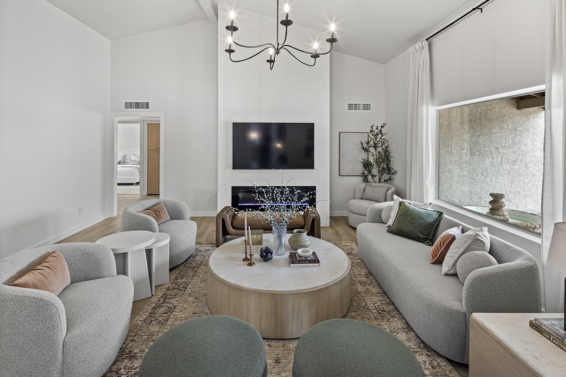 Modern living room with white walls, a black chandelier, a large wall-mounted TV, and a fireplace below. There are multiple light gray sofas and chairs arranged around a large round white marble coffee table with decorative items and books. A large window with white curtains and a small indoor plant add natural light and greenery.