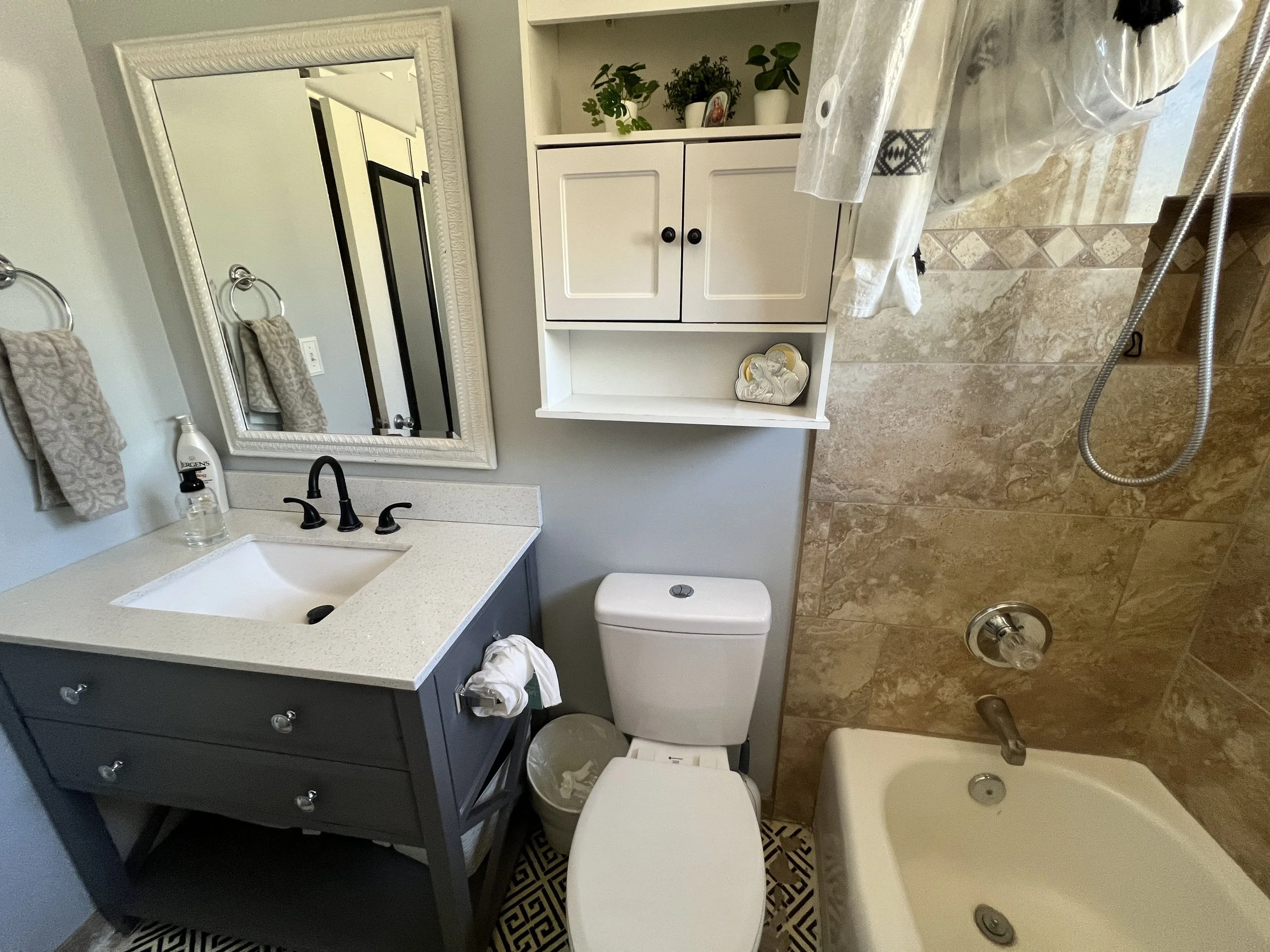 Bathroom renovation featuring wood vanity with brass fixtures, floral wallpaper, beadboard wainscoting, white toilet, and arched walk-in shower with gold shower system.