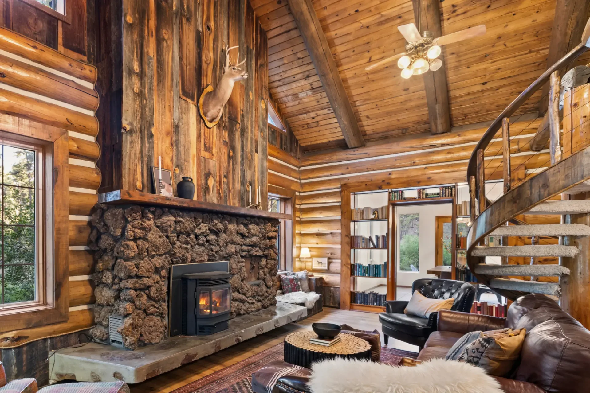 Rustic cabin interior featuring a stone fireplace, log walls, a spiral staircase, and a mounted deer head