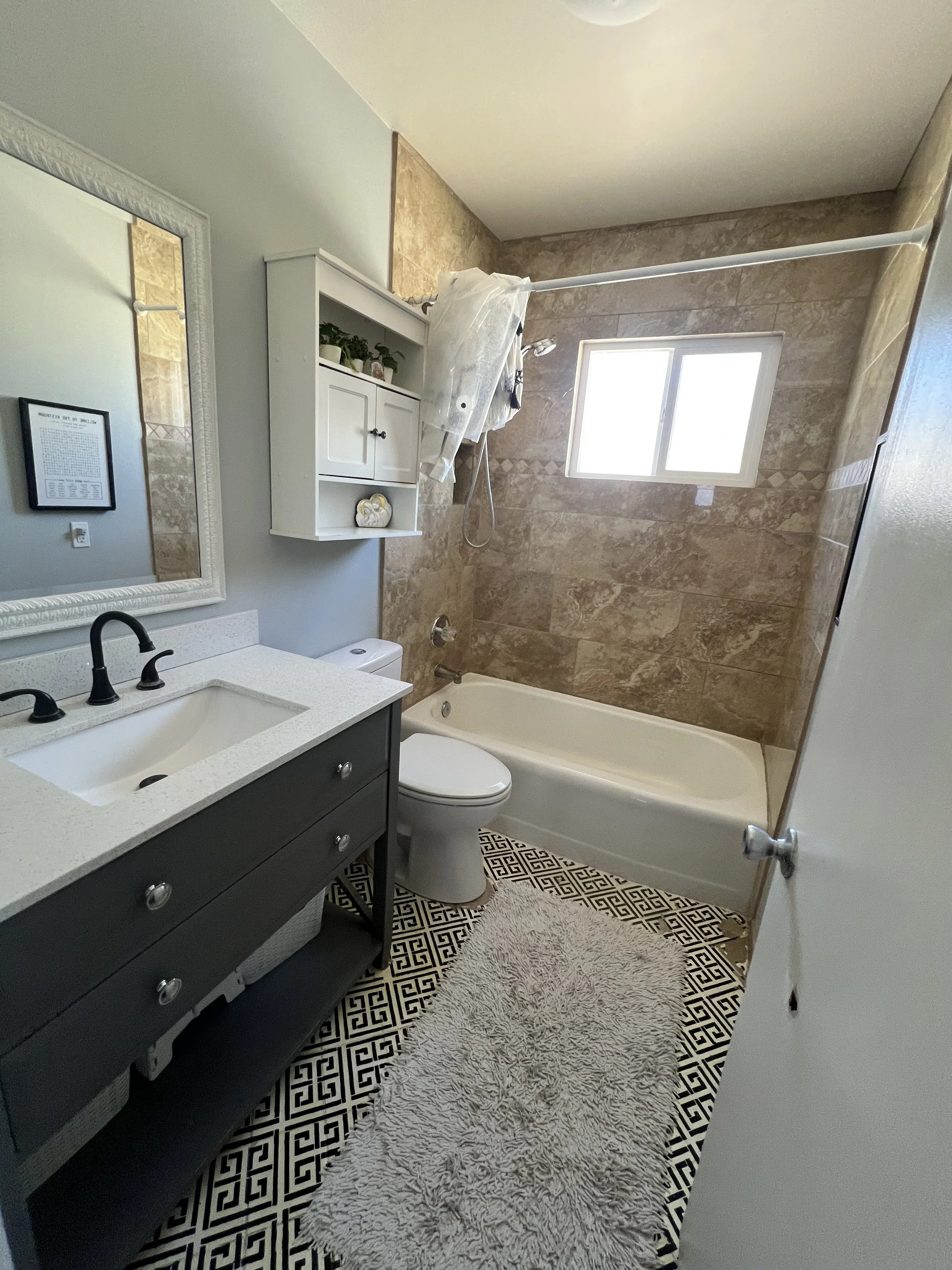 Bathroom renovation featuring wood vanity with brass fixtures, floral wallpaper, beadboard wainscoting, white toilet, and arched walk-in shower with gold shower system.