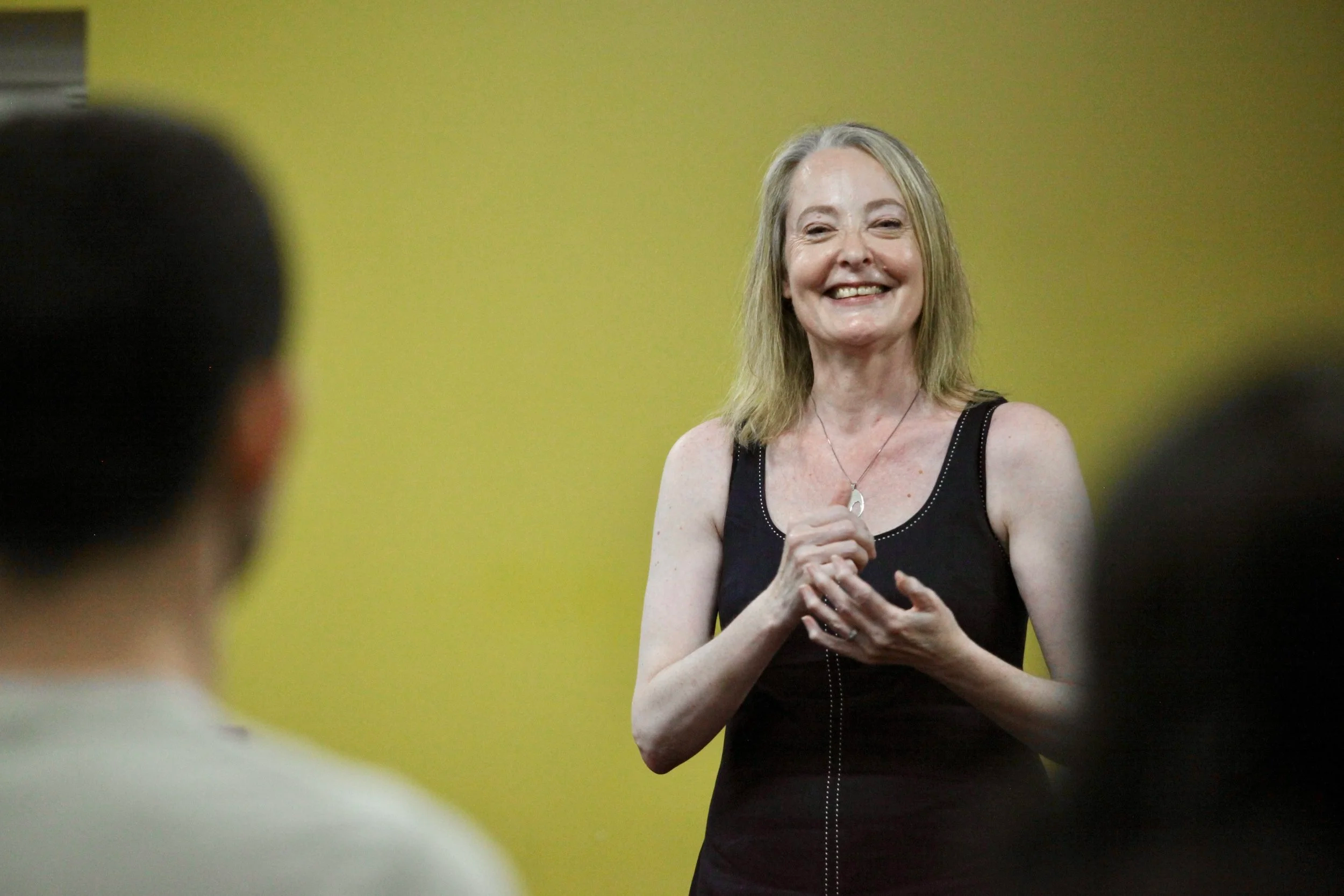 Sheena Pritchard is speaking on resilience& adversity.  She has shoulder-length blonde hair, is smiling and gesturing with her hands.  She is in front of a yellow wall.