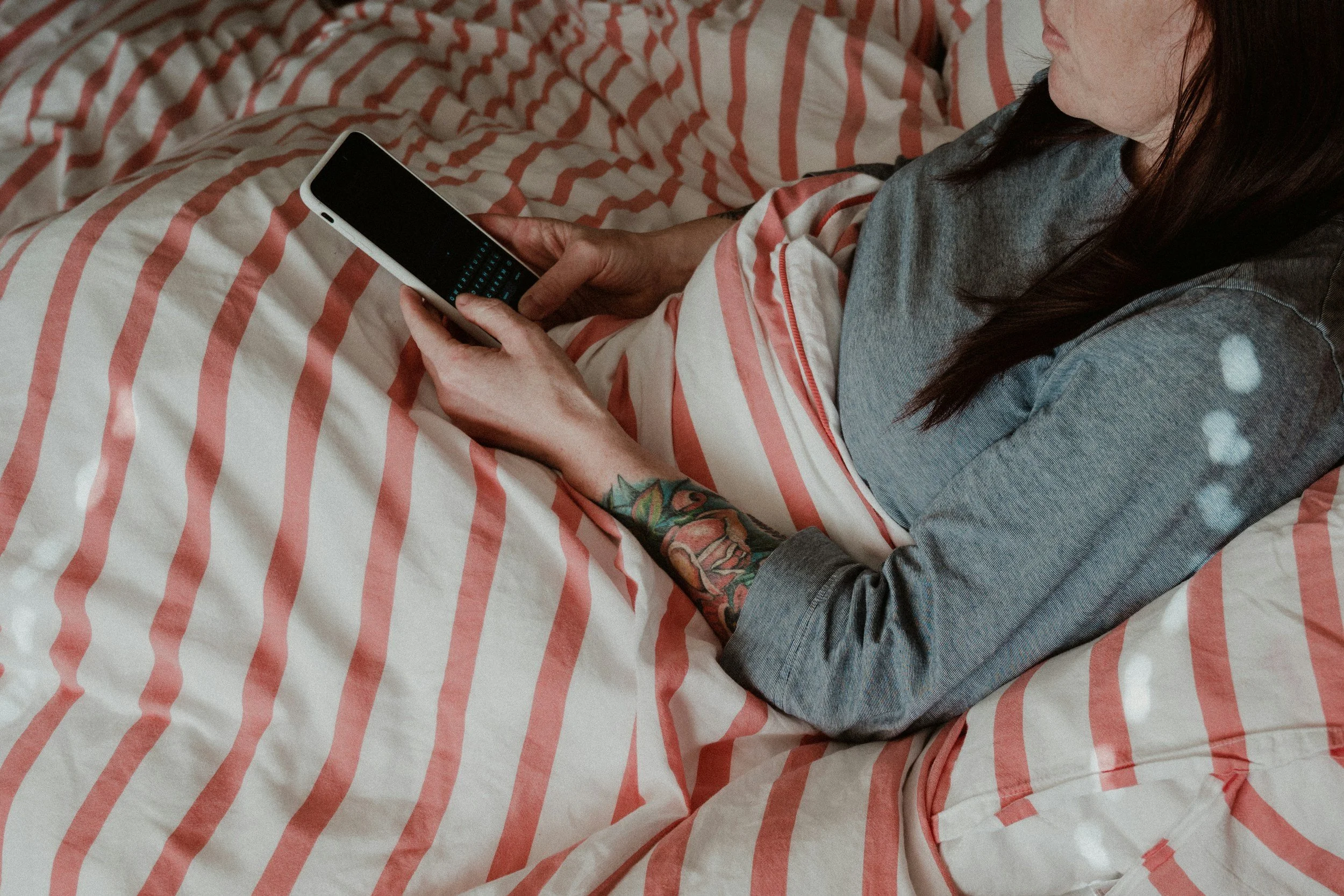 Person with chronic illness lying on bed with striped bedsheet, being coached via her smartphone, wearing a grey shirt, with dark hair and visible tattooed arm.