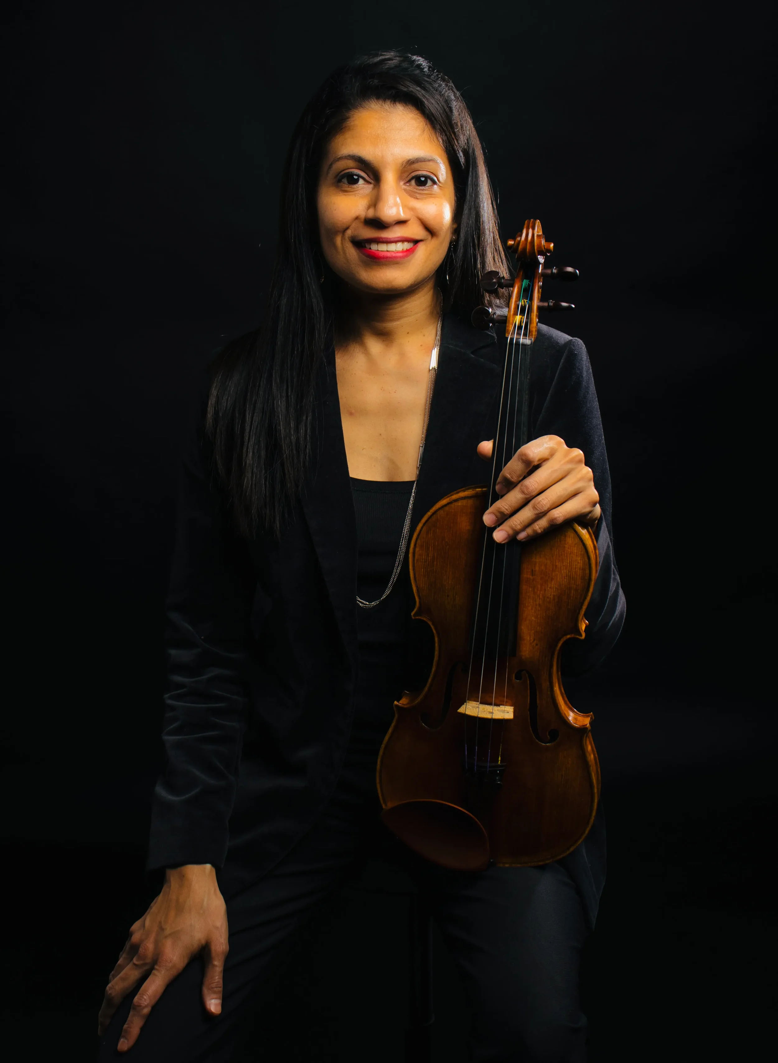 Woman holding a violin, dressed in black, against a dark background.