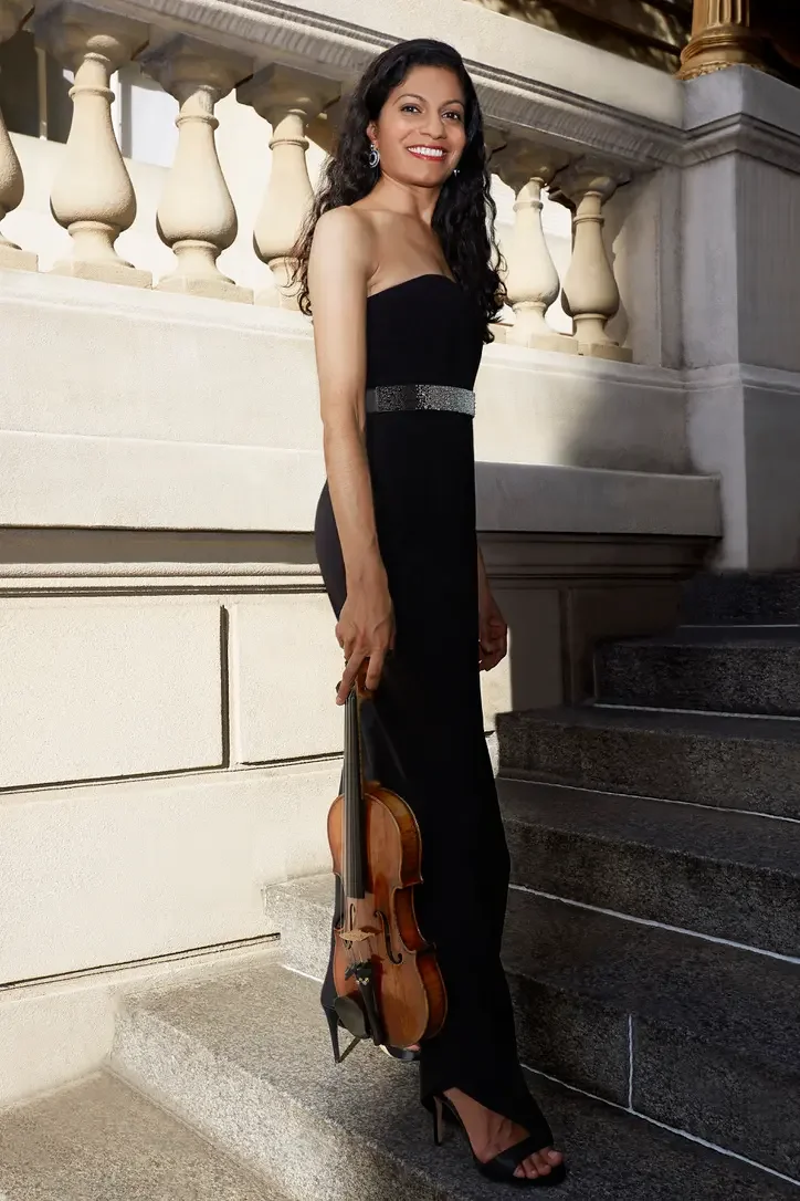 A woman in a strapless black evening gown holding a violin, standing on steps outside a grand building with stone balustrades and ornate columns, smiling at the camera.