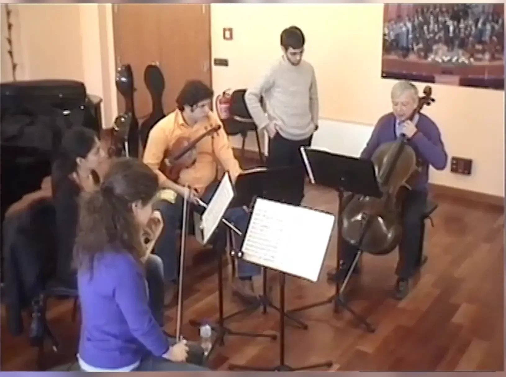 Group of six people in a room, five are playing string instruments, and one person is standing observing. The room has wooden floors, a fireplace, and a wall-mounted TV or art piece.