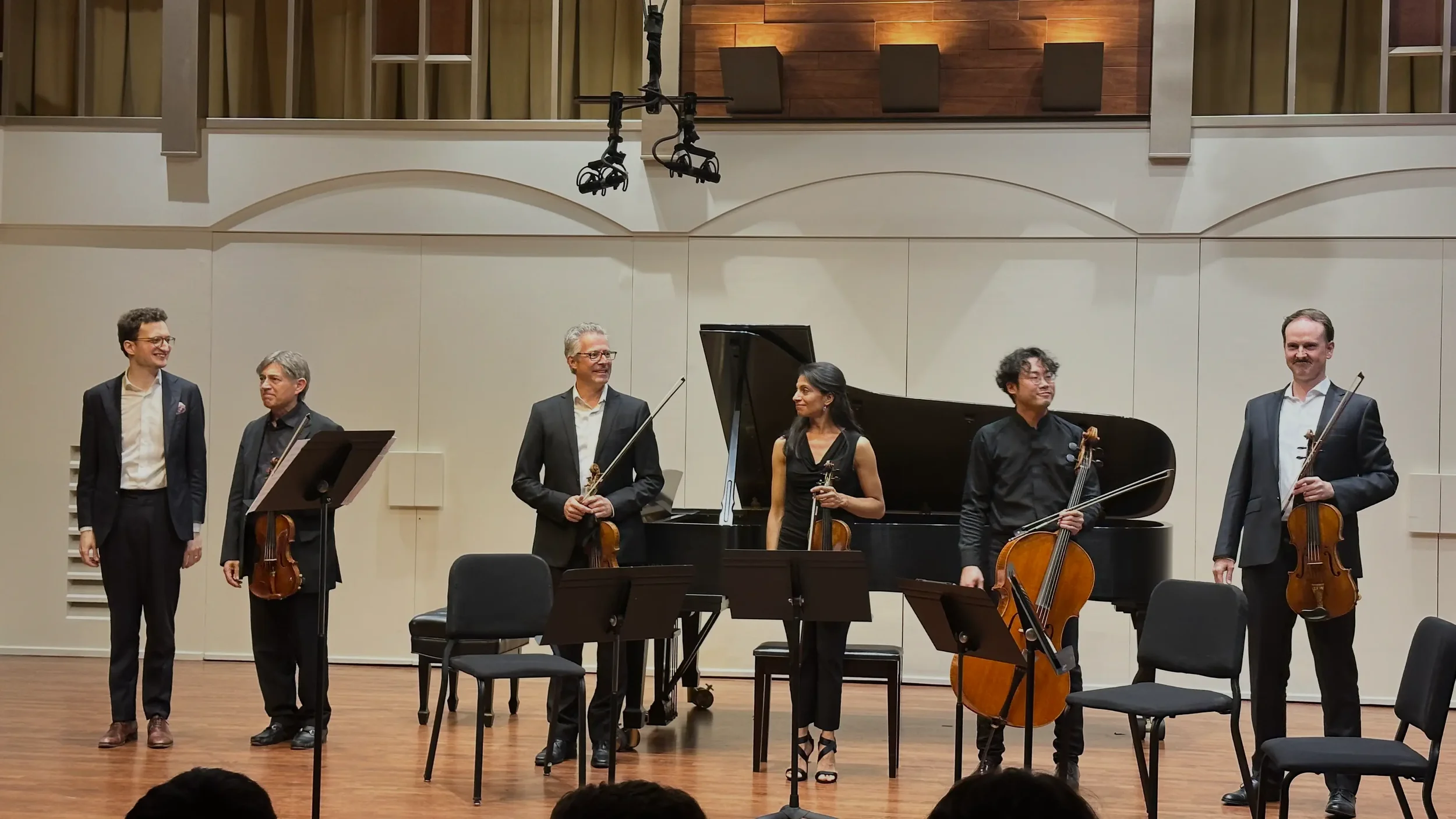 Six musicians standing on stage with string instruments in a concert hall, after a performance, with chairs and music stands in front of them.