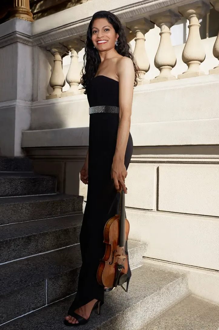 A woman in a strapless black evening gown holding a violin while standing on staircase outside of a classical building with columns and balustrade, smiling at the camera.