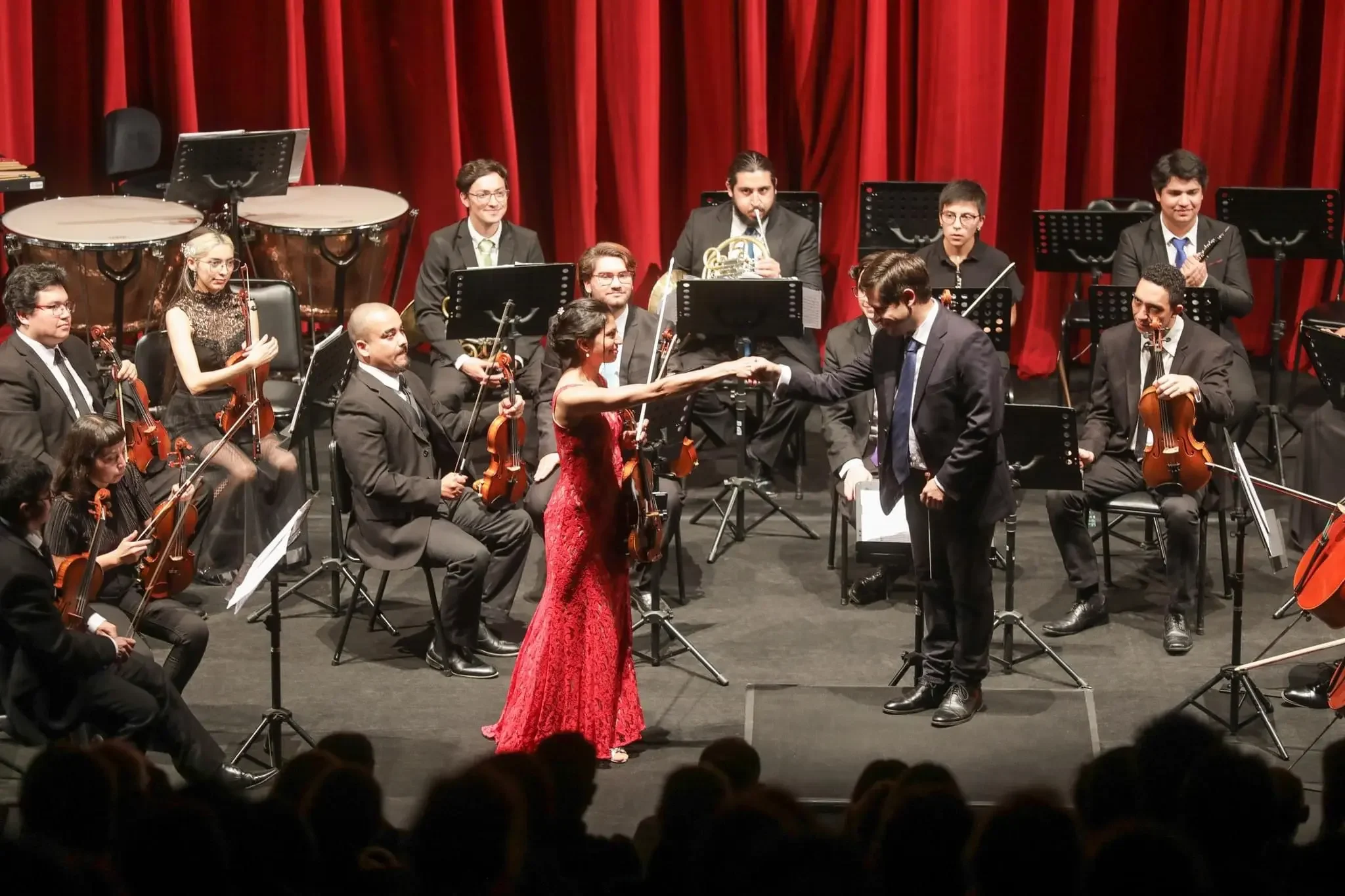 Orchestra on stage in concert hall with red curtains, woman in red gown shaking hands with man in black suit, musicians playing string and wind instruments