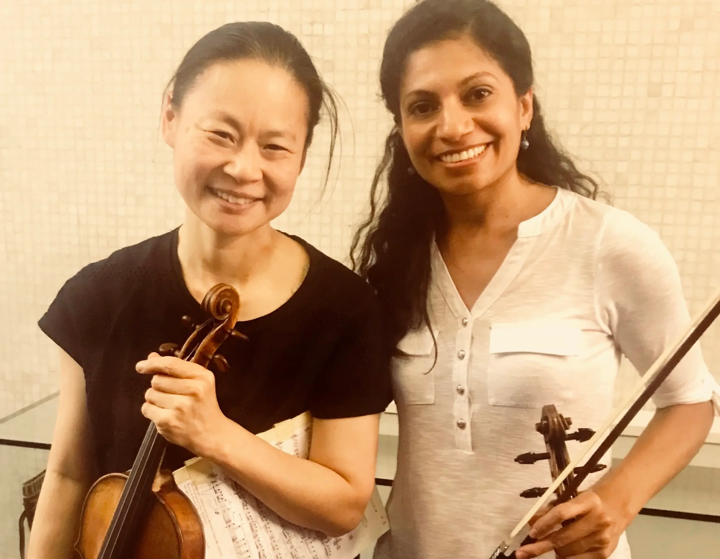 Two women, one Asian holding a violin, and one South Asian holding a violin and bow, smiling together indoors.
