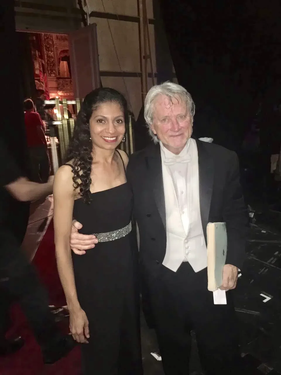 A woman in a black dress and a man in a tuxedo are standing together, smiling, at a formal event.