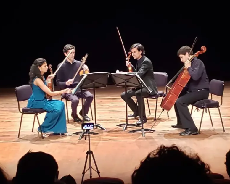 Four musicians performing a classical music concert on stage, including a violinist, a cellist, a pianist, and a violinist, with audience in front.
