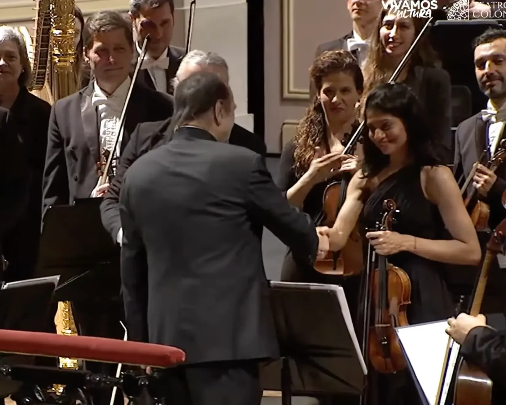 Orchestra members shaking hands with a conductor during a performance, some musicians holding violins and bows, in a concert hall setting.