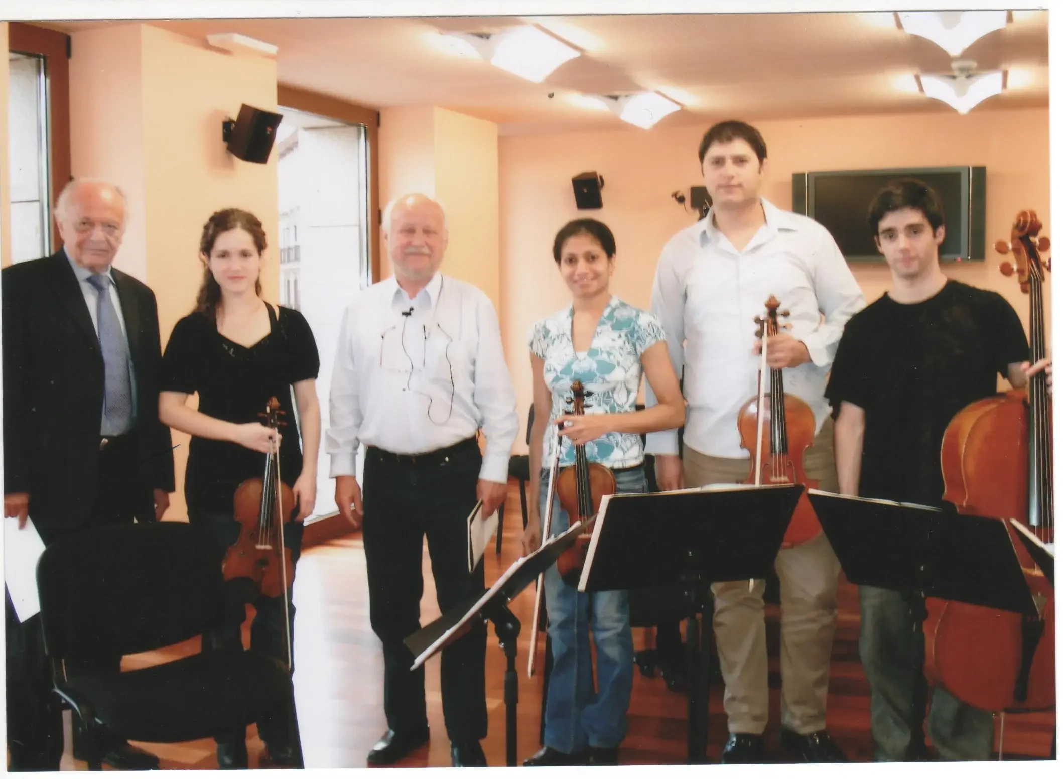 Six people standing in a room with musical stands and violins, possibly a classical music rehearsal or concert setting.