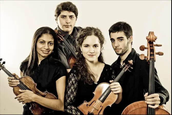 Four young musicians holding string instruments, two with violins and two with cellos, standing together against a plain background.