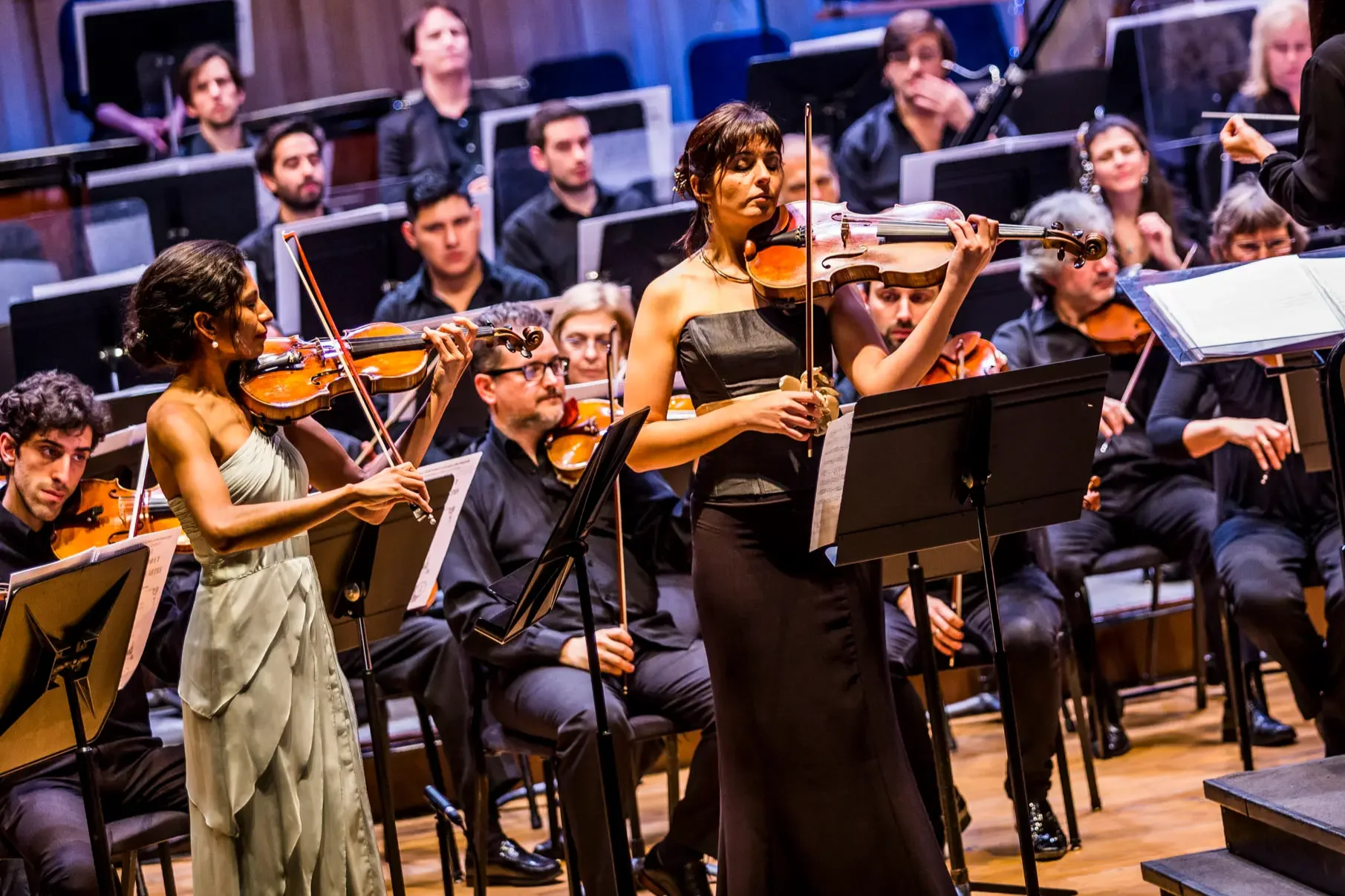 Orchestra performing on stage, with two violinists in the foreground, one in a white dress and the other in a black dress, while multiple other musicians are seated behind them.