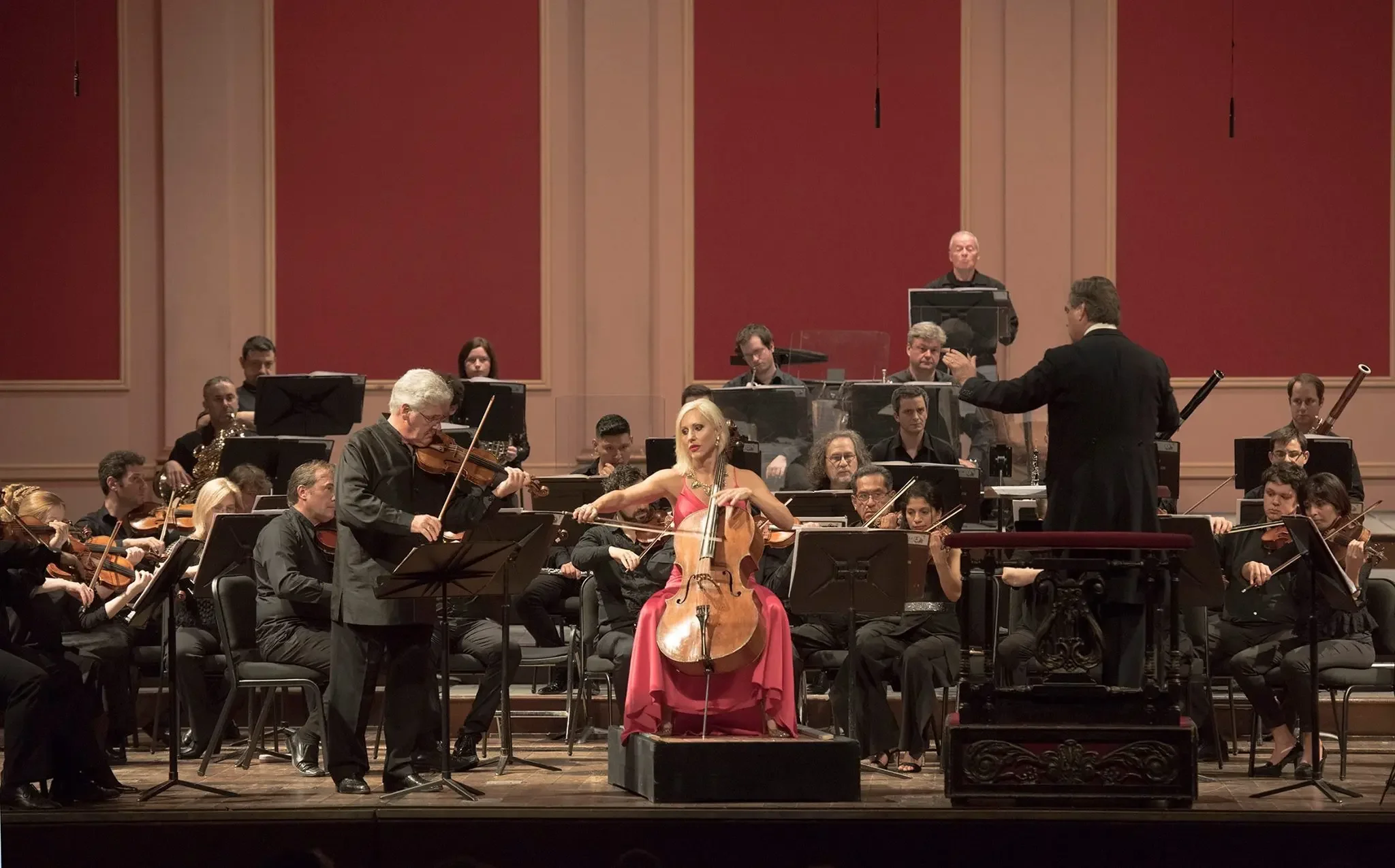 An orchestra performing on stage, with a female cellist in a red dress in the center, a violinist playing to her left, and a conductor directing the musicians. The orchestra members are seated with music stands, and the stage has a red and cream background.