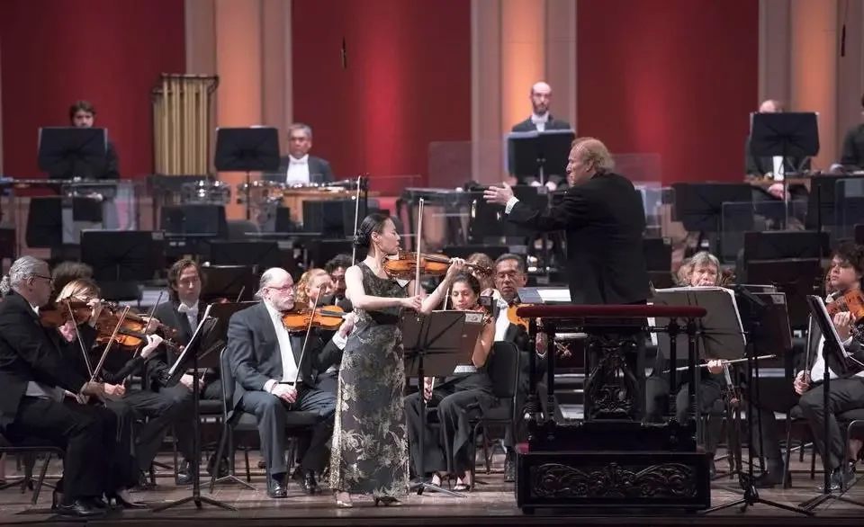 An orchestra performing on stage with a male conductor leading a female violinist, all dressed in formal black attire, with a red and orange lit background