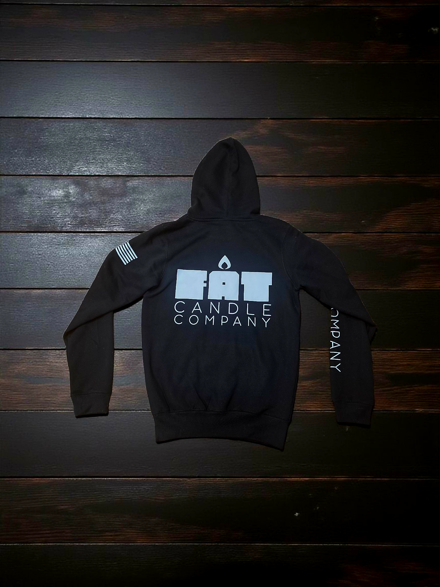 Black hoodie with the logo of Fat Candle Company, featuring a lit candle flame above the words, lying on a dark wooden surface.