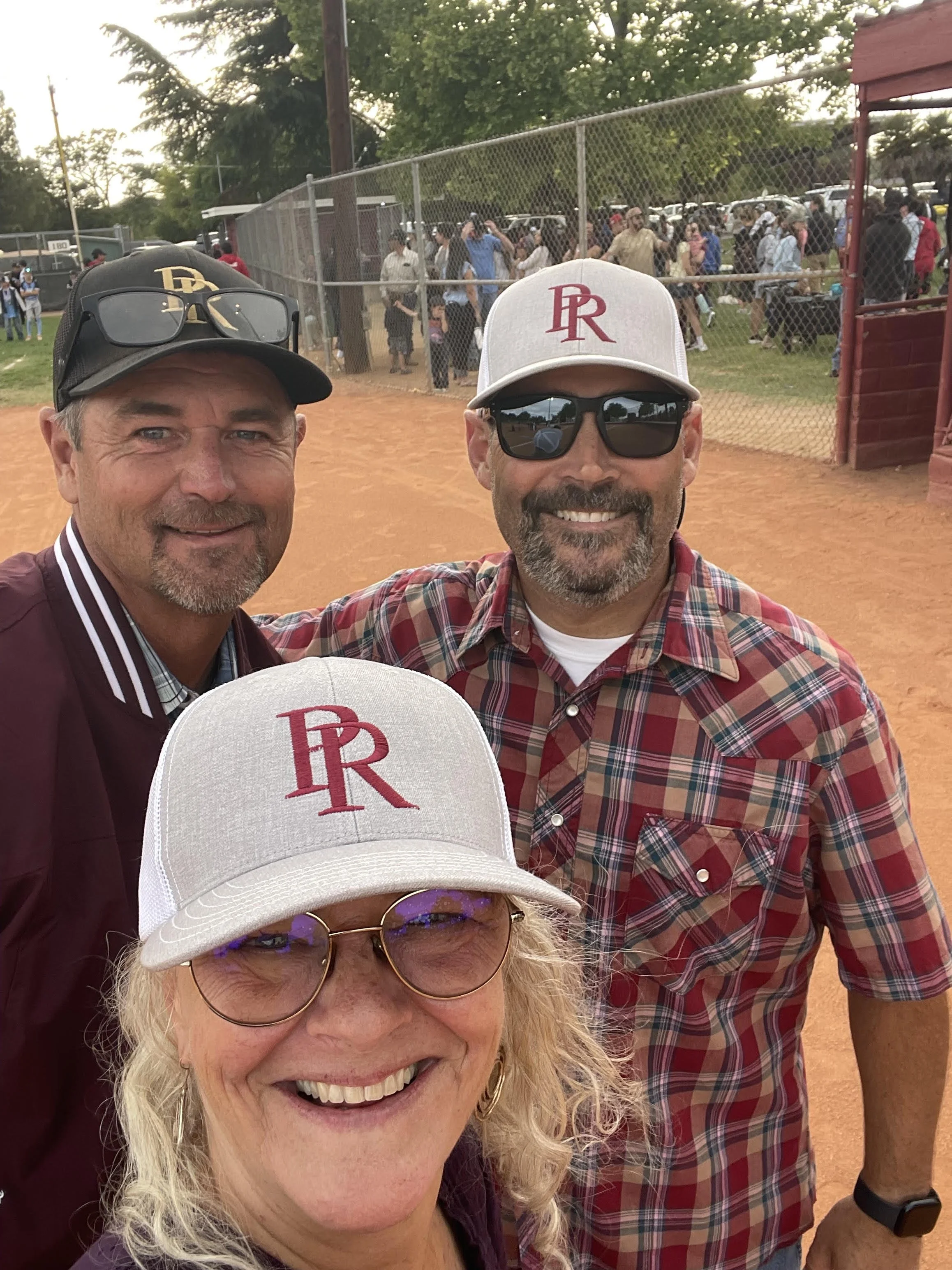 Three people taking a selfie at a baseball field, with a crowd of people and a chain-link fence in the background.