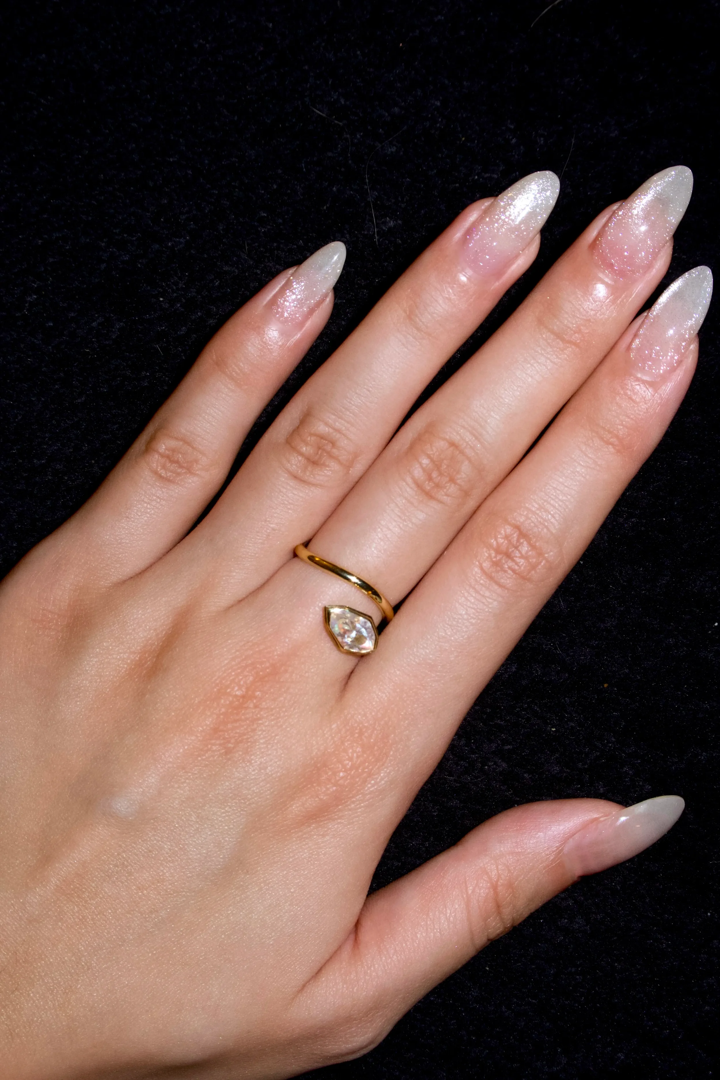 Close-up of a hand with glittery, natural-style nails, wearing a gold ring with a large, teardrop-shaped clear gemstone on the ring finger, against a black background.
