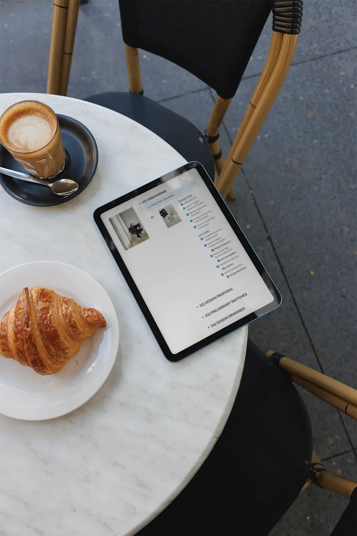 A marble table with a laptop displaying a project outline, a plate with a croissant, and a glass of coffee with milk on a saucer with a spoon. There is a black chair with wood arms nearby, on a dark outdoor surface.