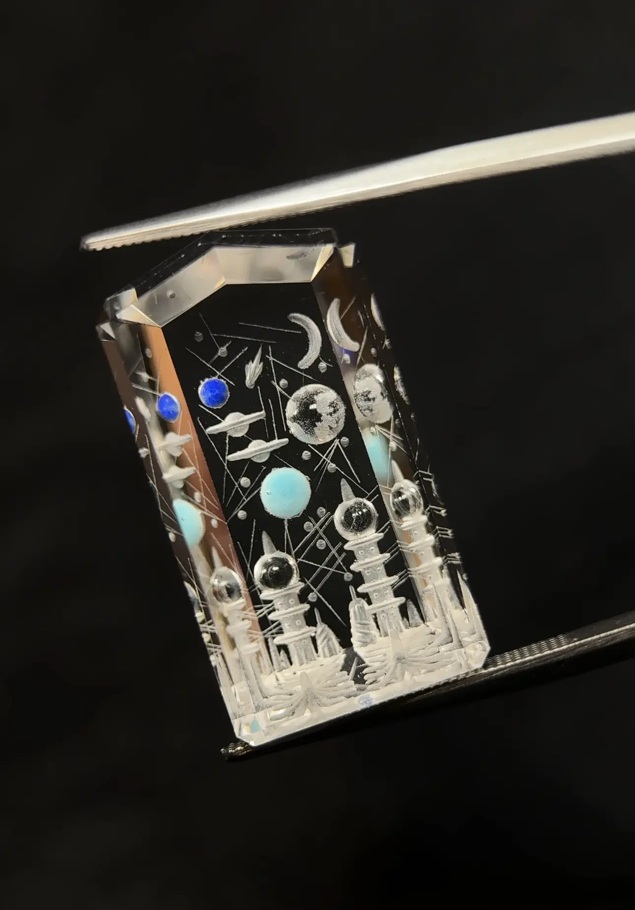 A clear rectangular glass or crystal piece engraved with cosmic-themed symbols, planets, and celestial objects, resting on a metallic tool or stand against a black background.