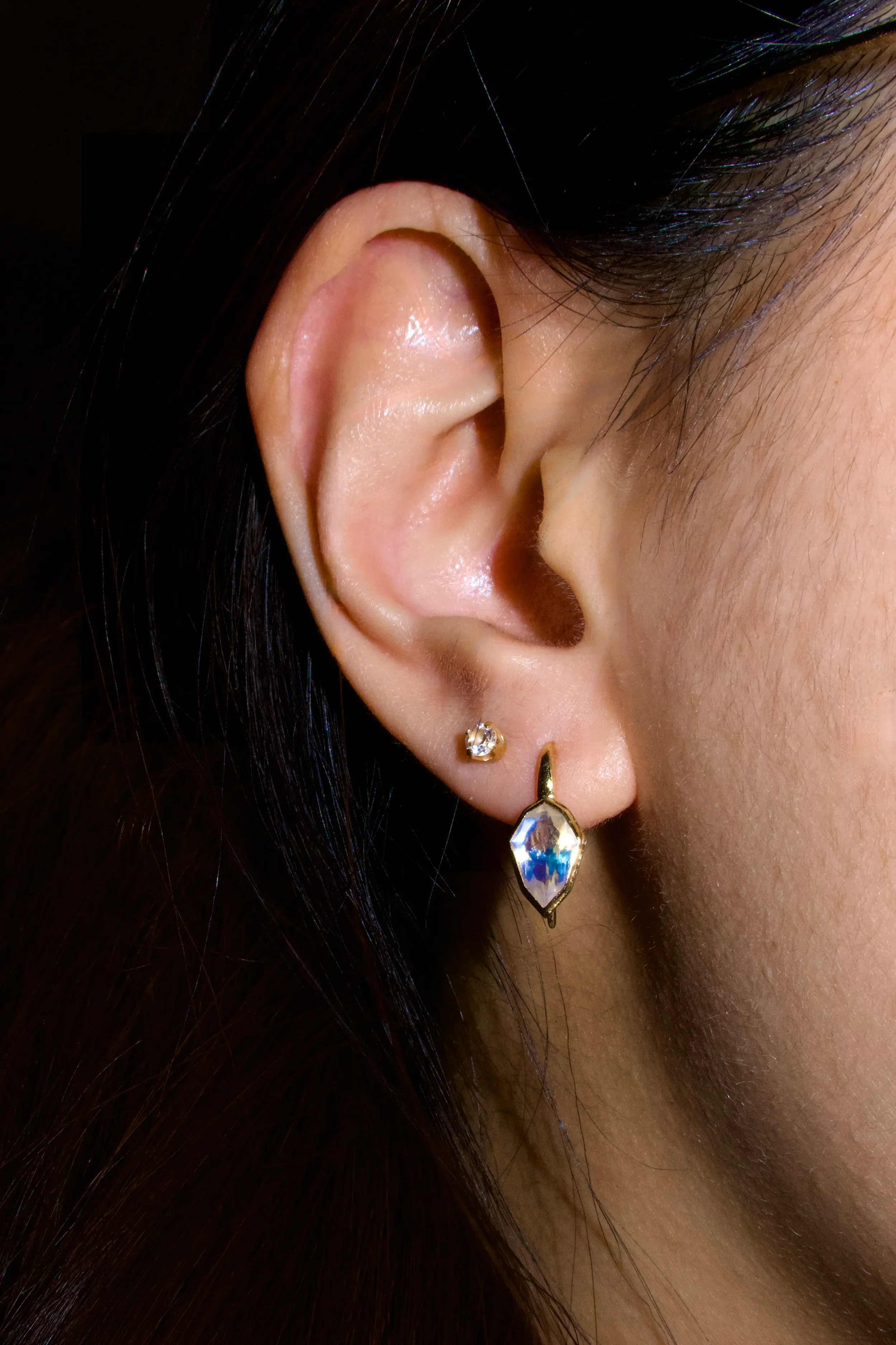 Close-up of a woman's right ear with two earrings: a small diamond stud and a larger teardrop-shaped blue gemstone earring with a gold setting.