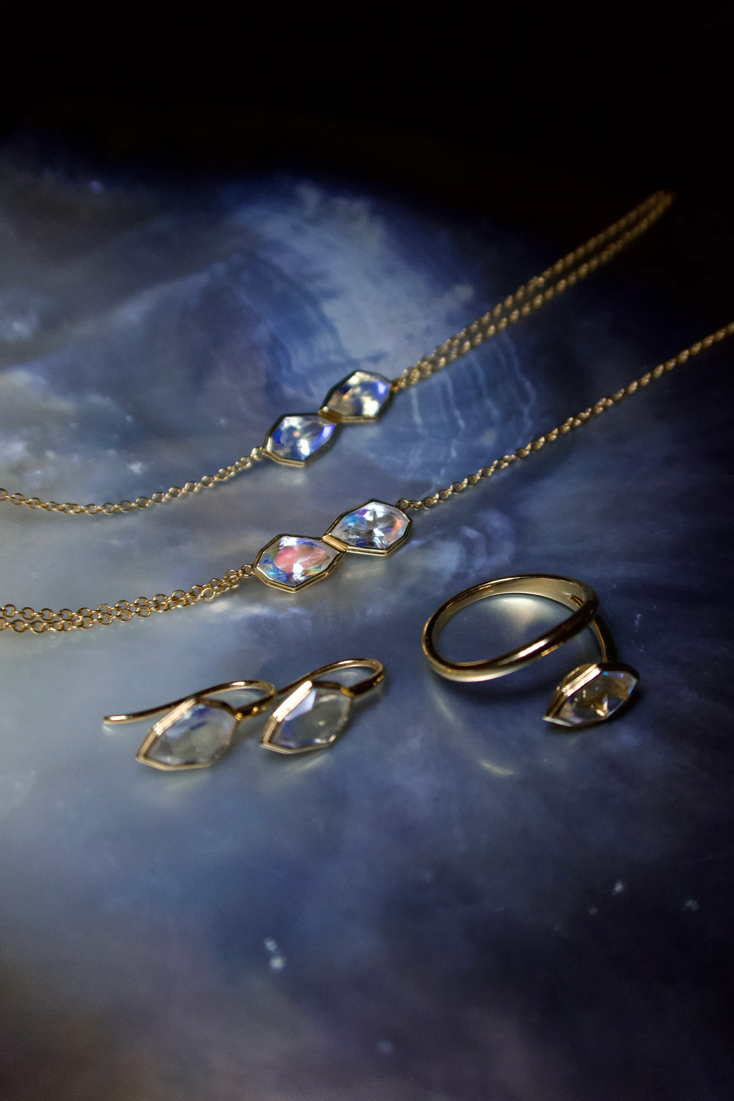 Set of gold jewelry including two necklaces, a ring, and earrings, all featuring large clear gemstones, resting on a dark, reflective surface.