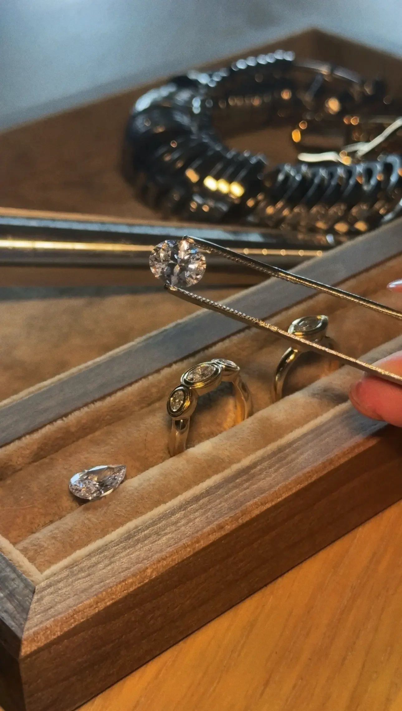 A jewelry box containing rings, a diamond, and a bracelet, with a hand using tweezers to hold a diamond for setting.