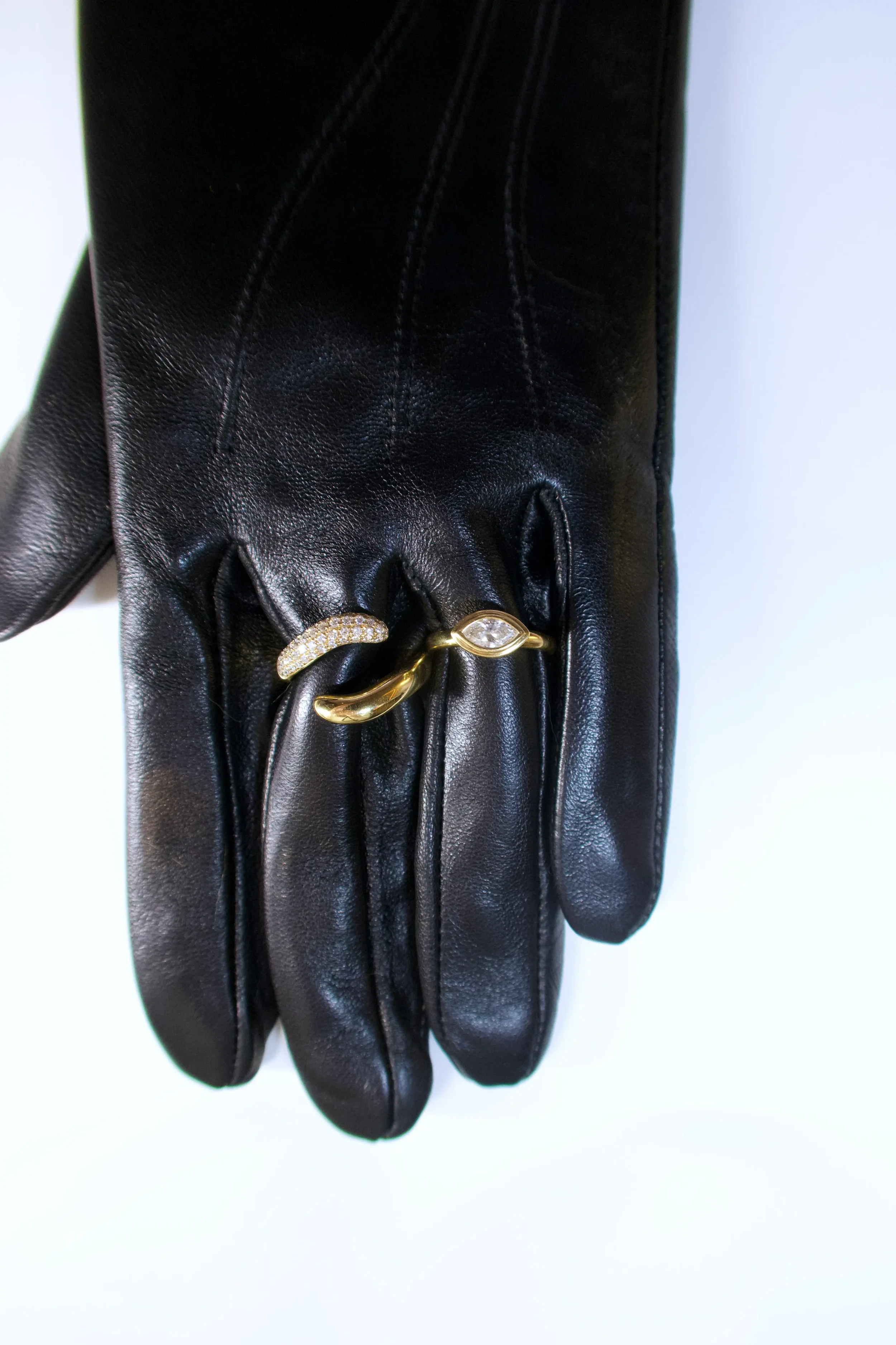 A black leather glove with three rings on it, including a gold band with a diamond, a gold ring with a curved design and a row of small diamonds, and a gold ring with a marquise-shaped diamond.