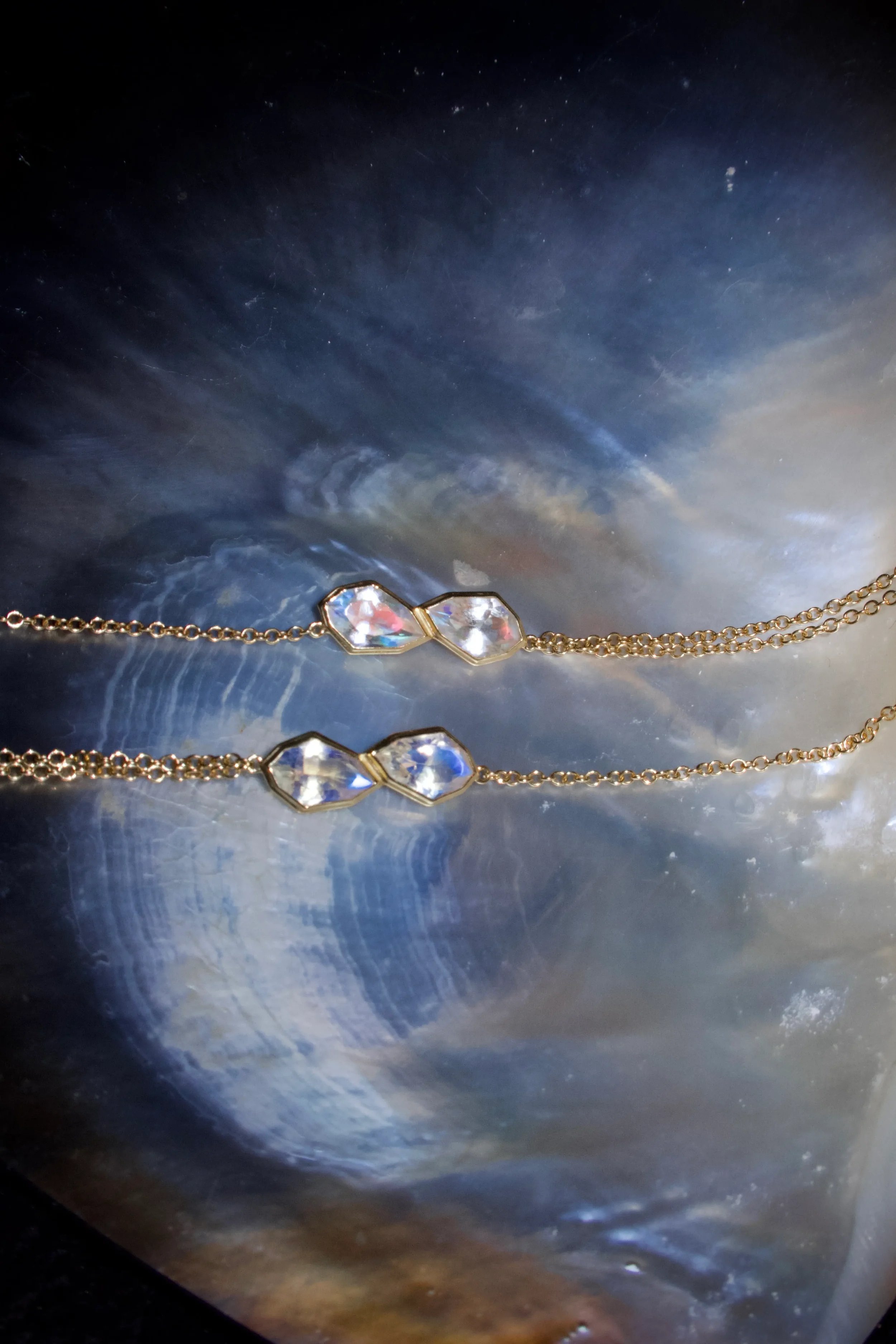 Two delicate gold chain necklaces with geometric-shaped, iridescent beads rest on a shiny, dark, swirling shell surface.