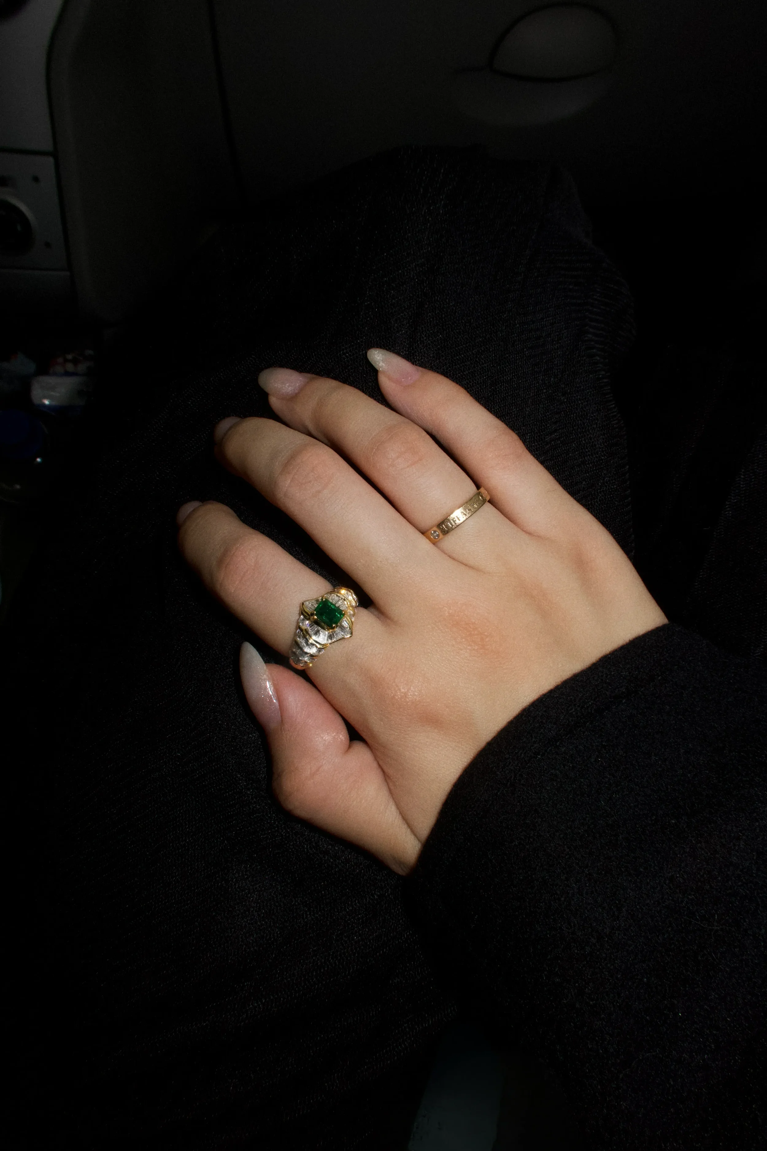 A person's hand displays two rings: one with a green gemstone and smaller diamonds, and a plain gold band, resting on a person's lap dressed in black pants.
