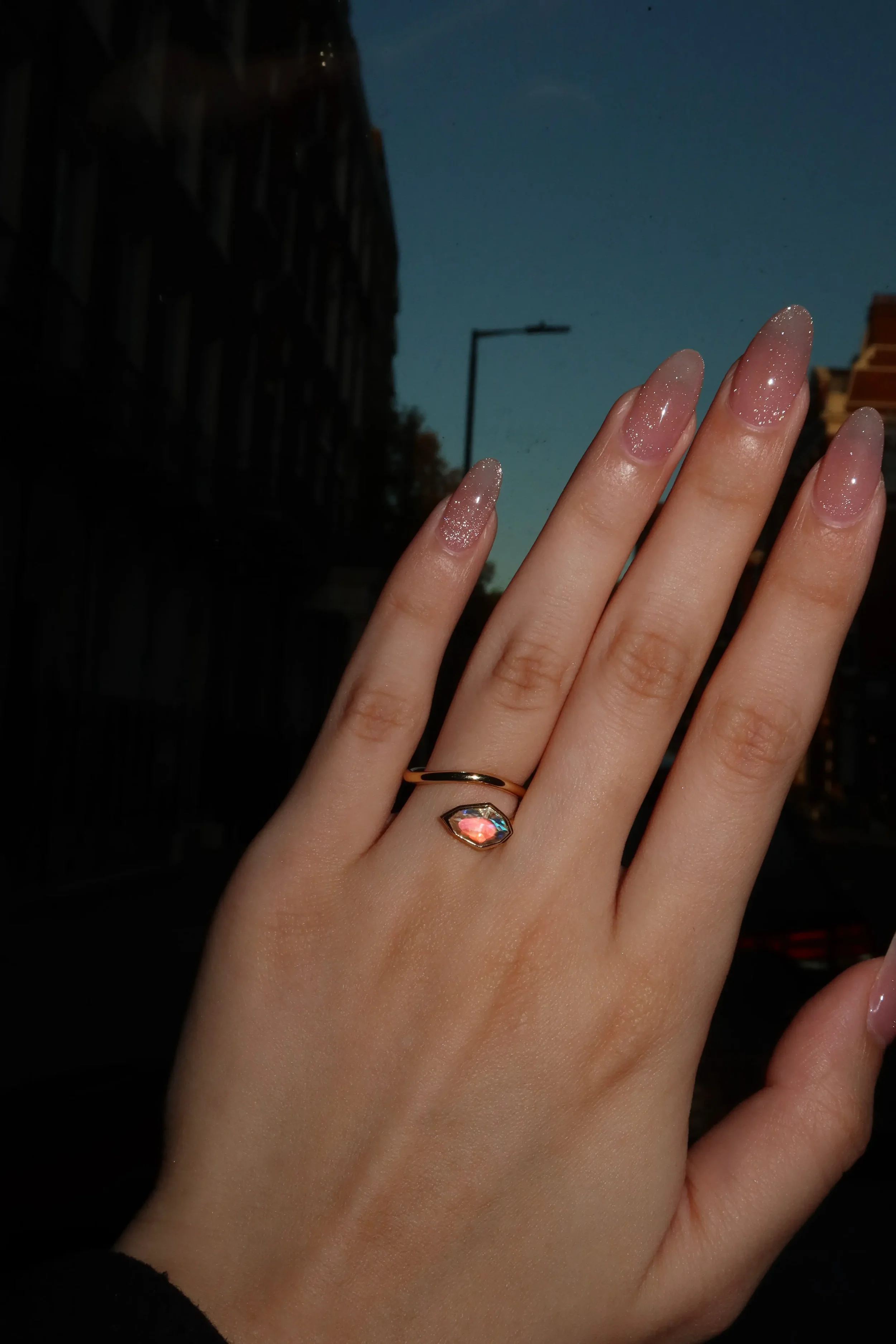 A hand with sparkly pink gel nails wears a gold ring with a pink gemstone, held up against a dark city street background at dusk.