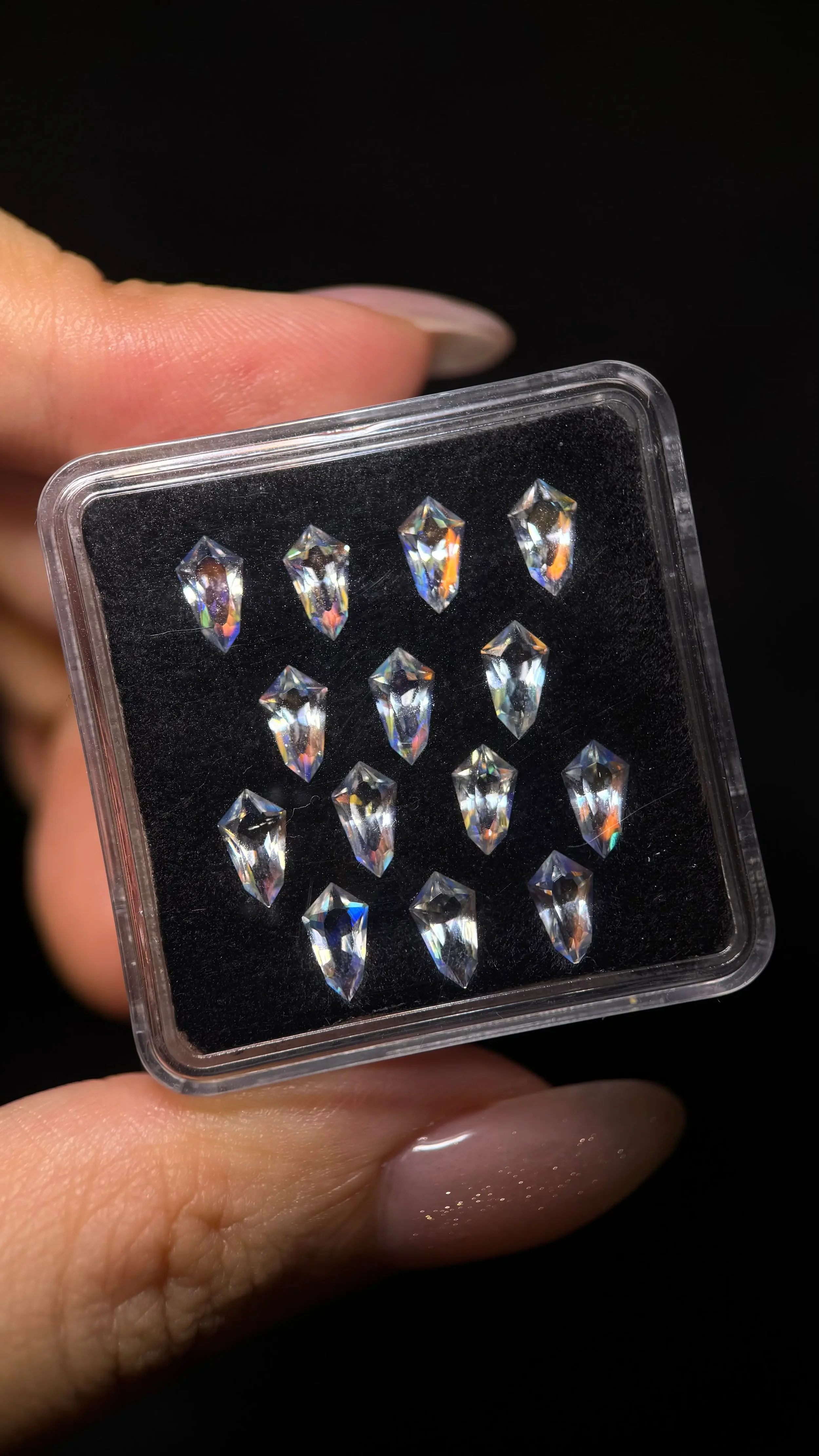 A hand holding a small plastic case with multiple small, clear, faceted gemstones or rhinestones inside.