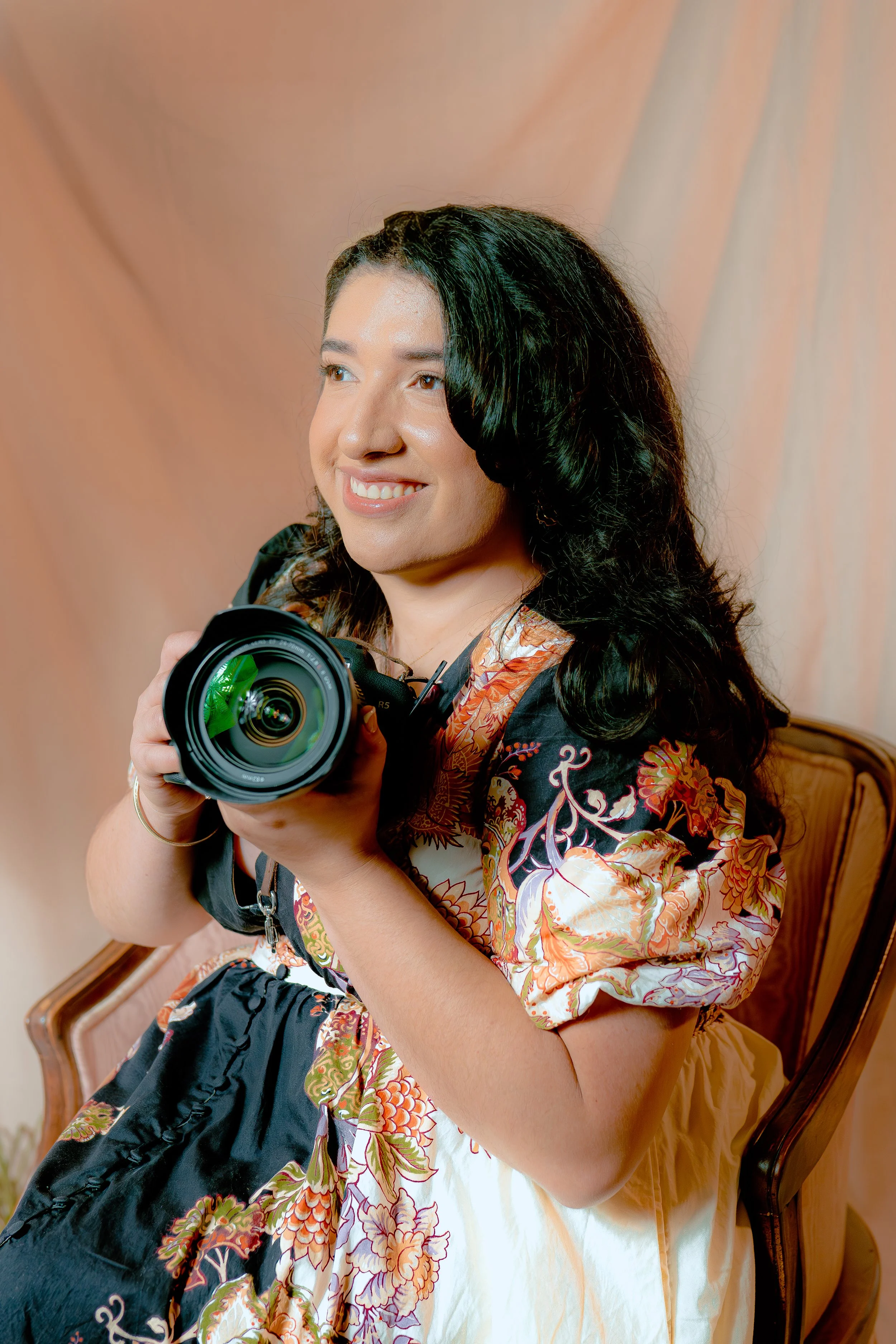 Woman with black, curly hair smiling and holding a camera while sitting on a brown wooden chair with a beige curtain background.