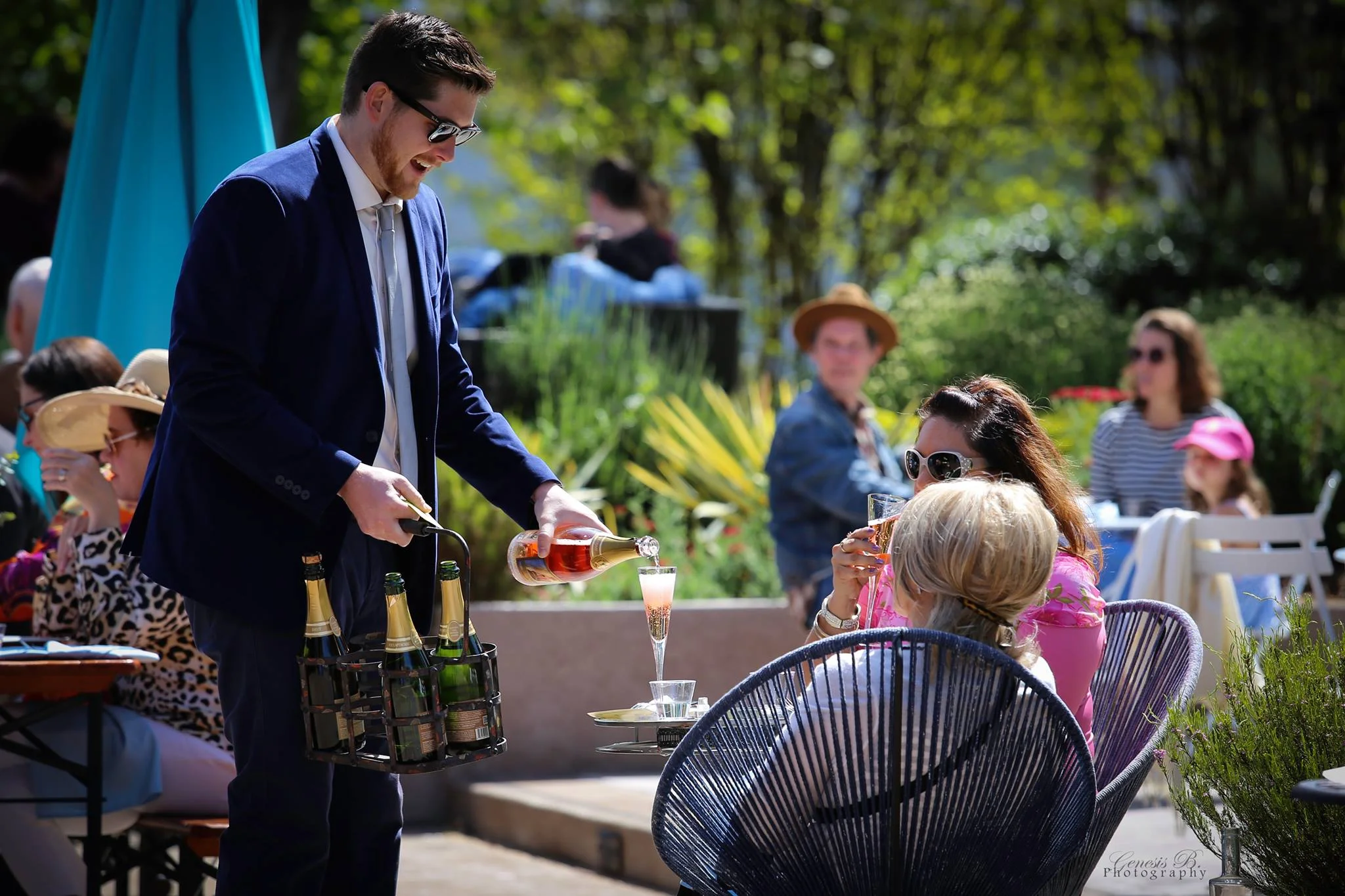 A man in a navy suit pouring champagne into a glass for a woman wearing sunglasses at an outdoor gathering.