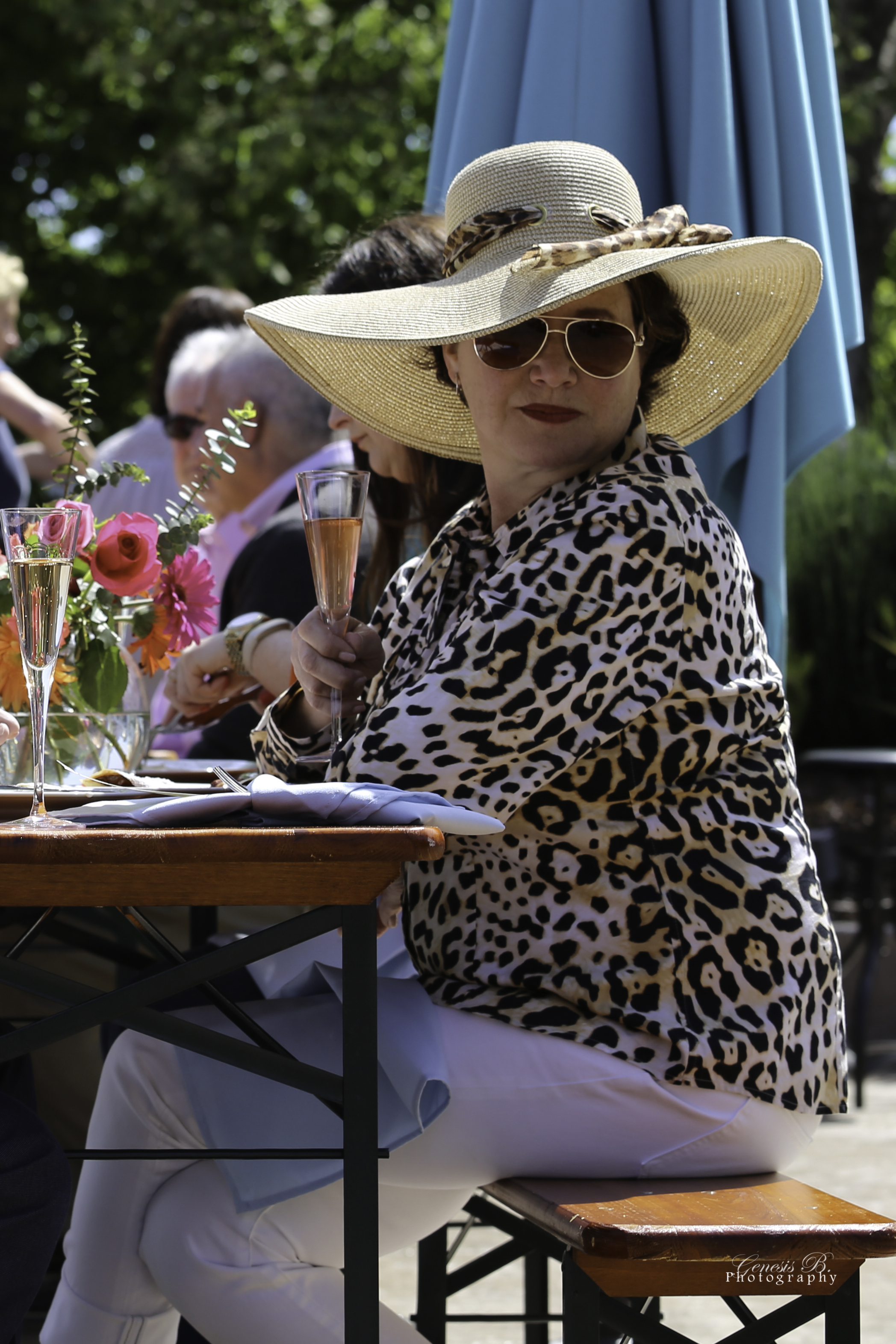 A woman wearing a large straw hat, sunglasses, a leopard print shirt, and white pants, sitting at an outdoor table, holding a glass of pink champagne.