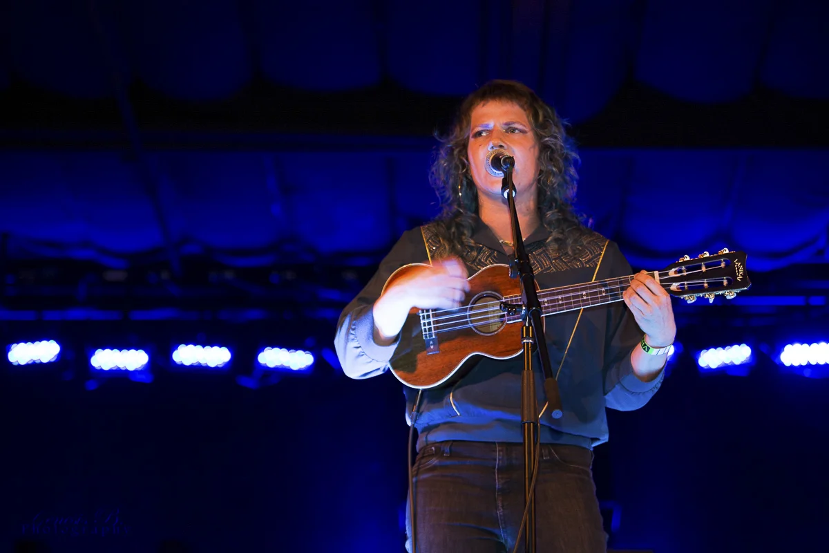 A person with curly hair singing into a microphone while playing a ukulele on stage with blue lighting.