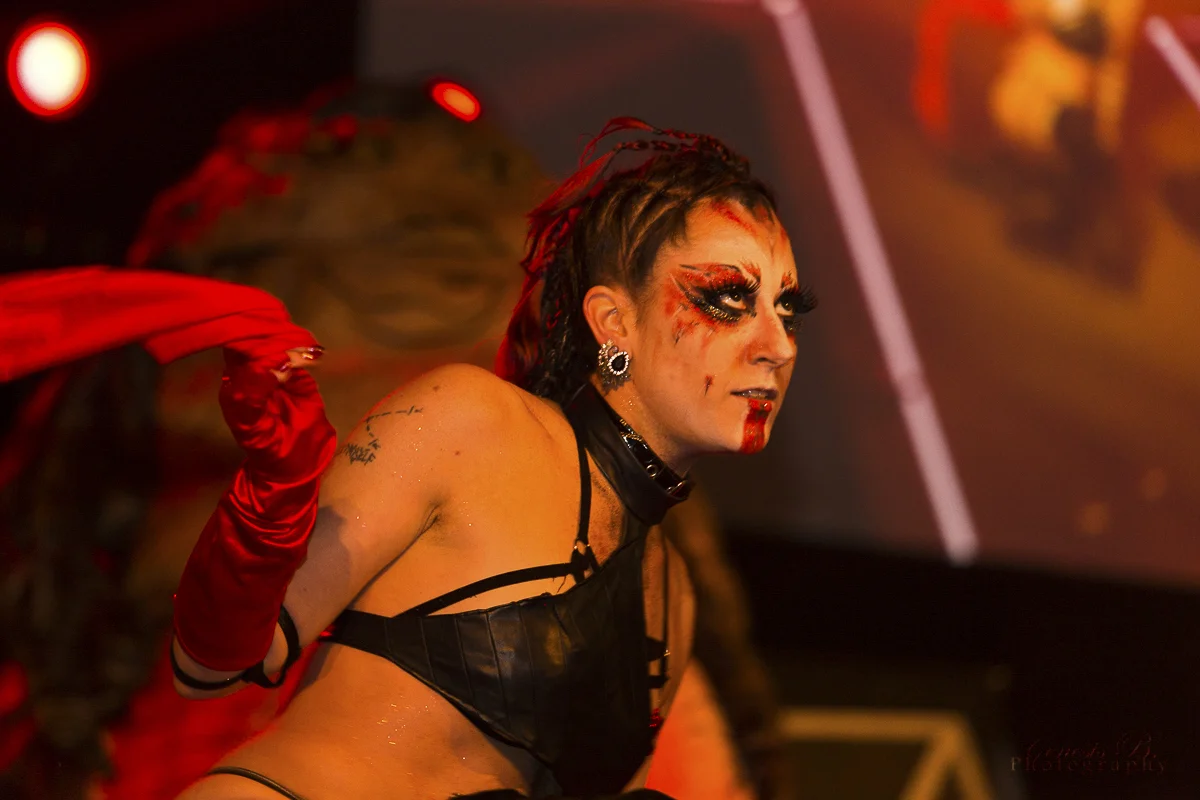 A person with light skin, elaborate makeup, and styled hair, wearing a revealing black leather outfit, red gloves, and dramatic makeup with face paint, in a dimly lit setting with red lighting.