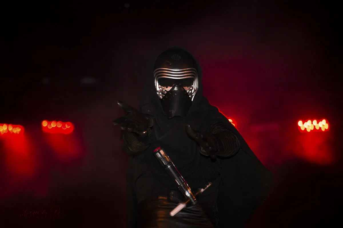 A person wearing black clothing, gloves, a mask, and a helmet with illuminated patterns, posing in a dark setting with red lights in the background.