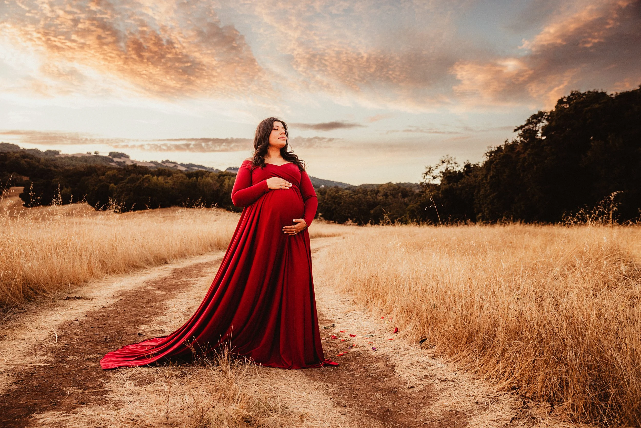 A pregnant woman in a long red dress standing on a dirt path in a field at sunset.